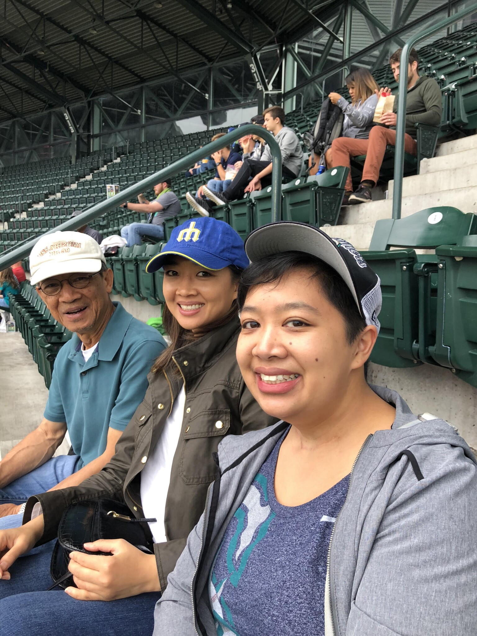 Three people sit smiling in a stadium with empty green seats around them. Two women and one man are wearing casual clothes and caps, with other spectators visible in the background.