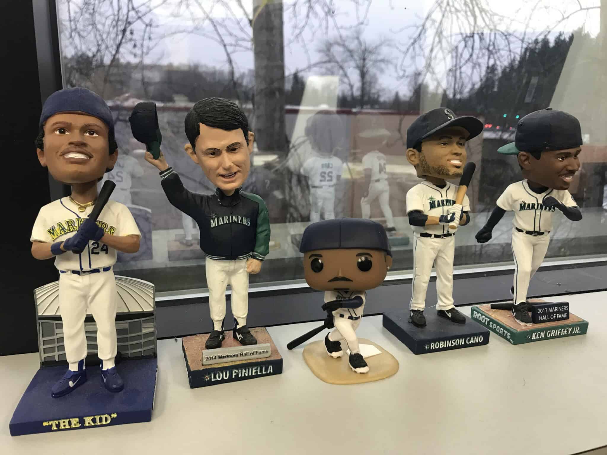 Five Seattle Mariners baseball player bobbleheads, including players and a manager, are lined up on a windowsill with trees visible outside. The bobbleheads are in various uniforms and poses.