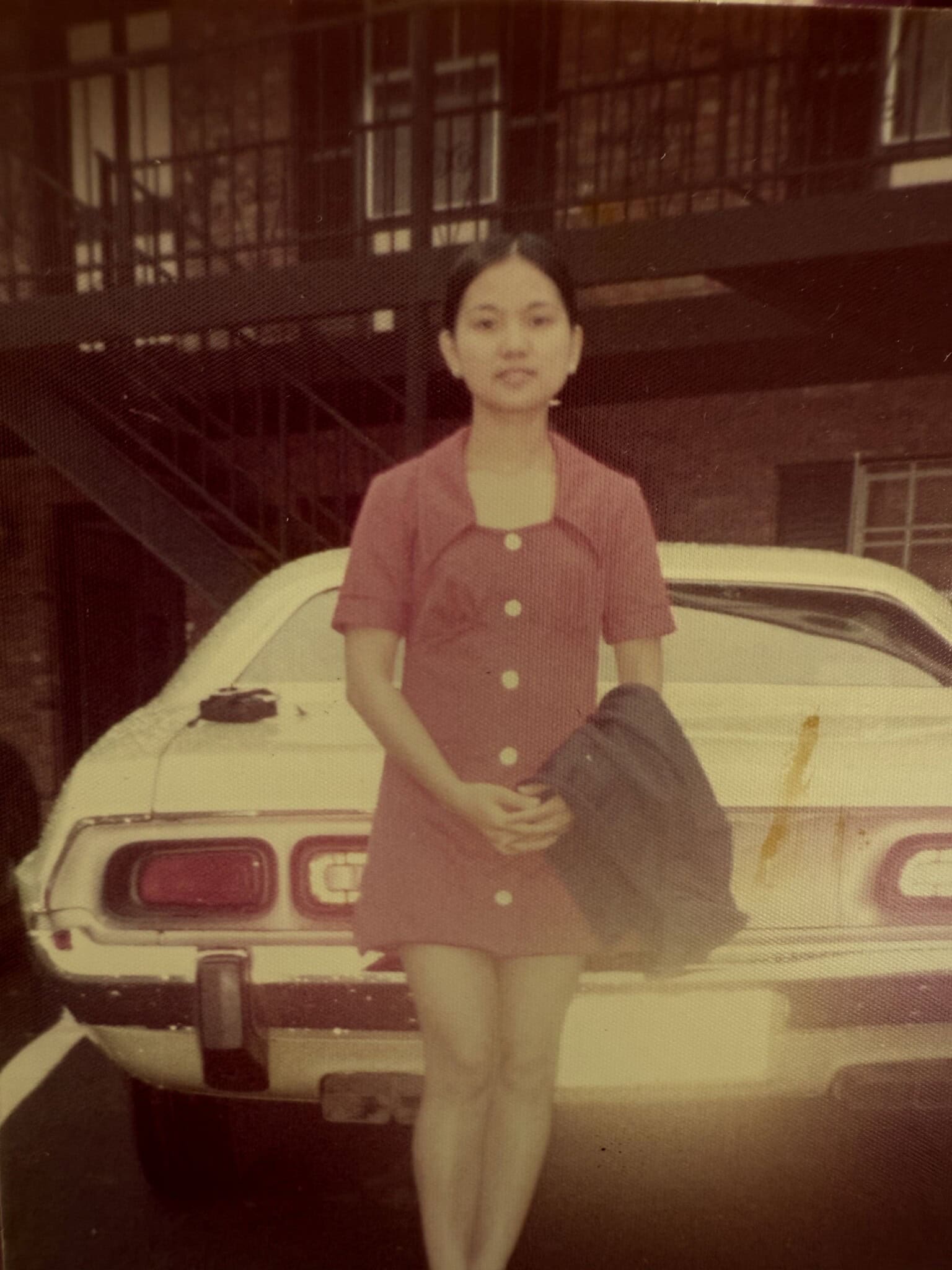 A young woman in a short-sleeved, button-up red dress stands in front of an old white car, holding a dark jacket—reminiscent of the style worn by early Filipino nurses. An outdoor staircase and brick building are visible in the background.