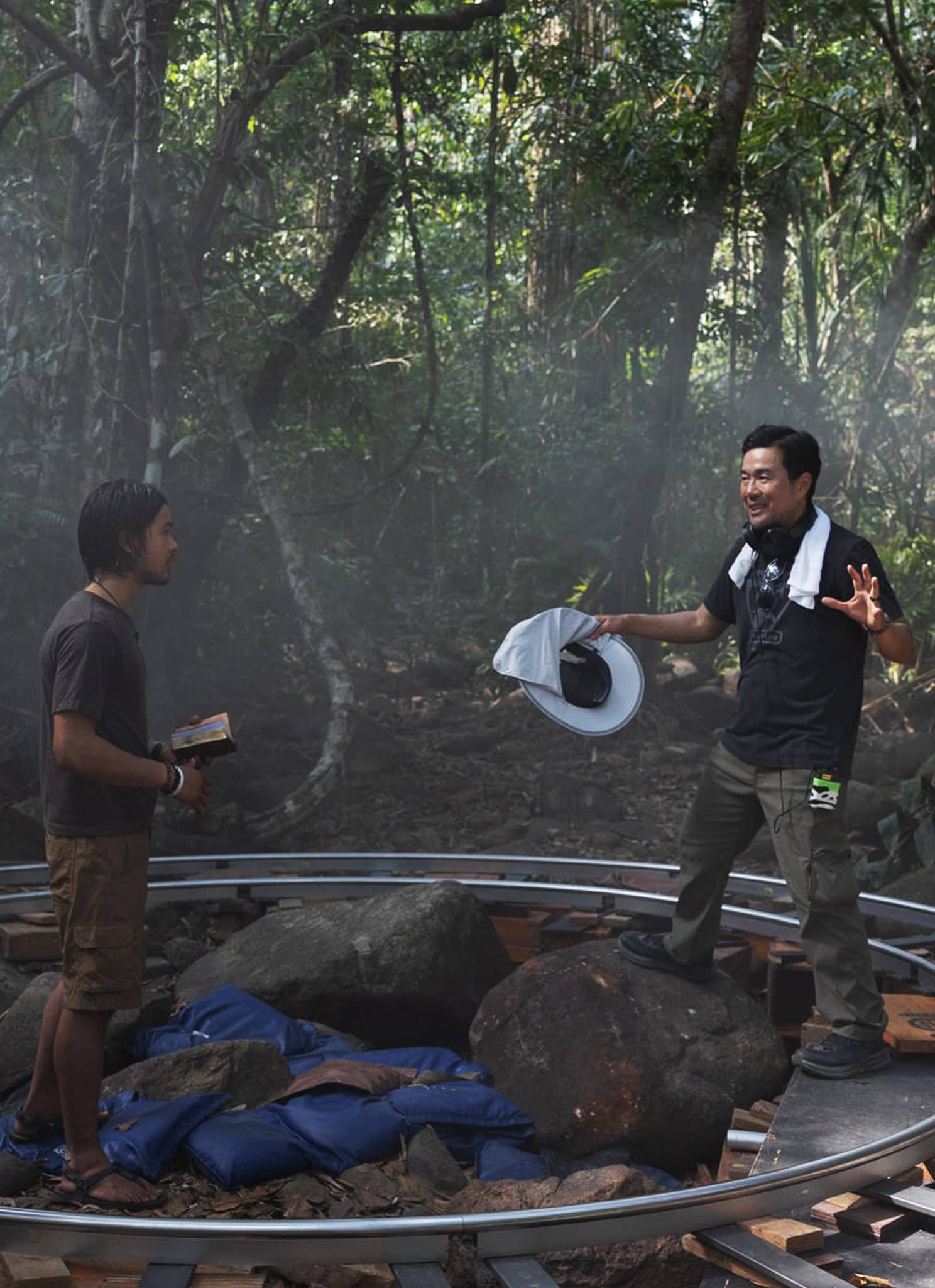 A film director gestures while talking to an actor on a forest set, surrounded by filming equipment and crew, with a camera on a track and sunlight filtering through the trees.