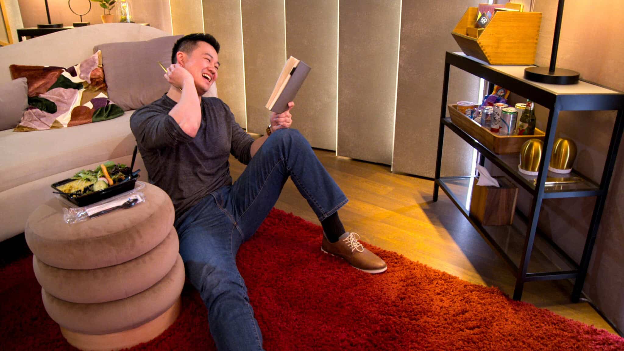 A man sits on a red rug, leaning against an ottoman, laughing while reading a book and chatting on the phone about Love Is Blind Season 9. A takeout meal sits nearby and the cozy, warmly lit room features a shelf stocked with drinks.