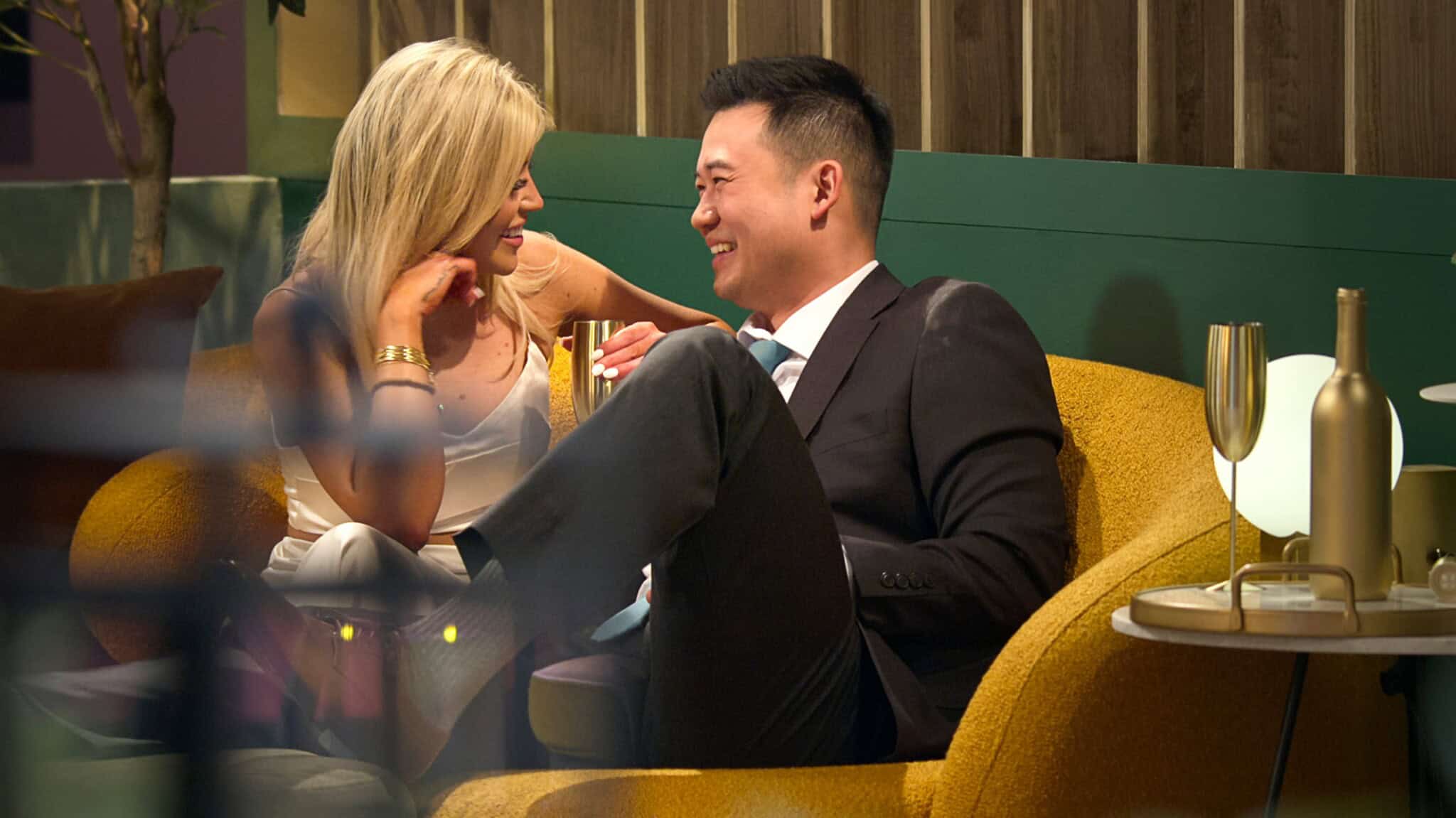 A woman in a white dress and a man in a suit sit close together on a yellow couch, smiling and looking into each other’s eyes. They appear to be enjoying a date, with drinks on the side table next to them.