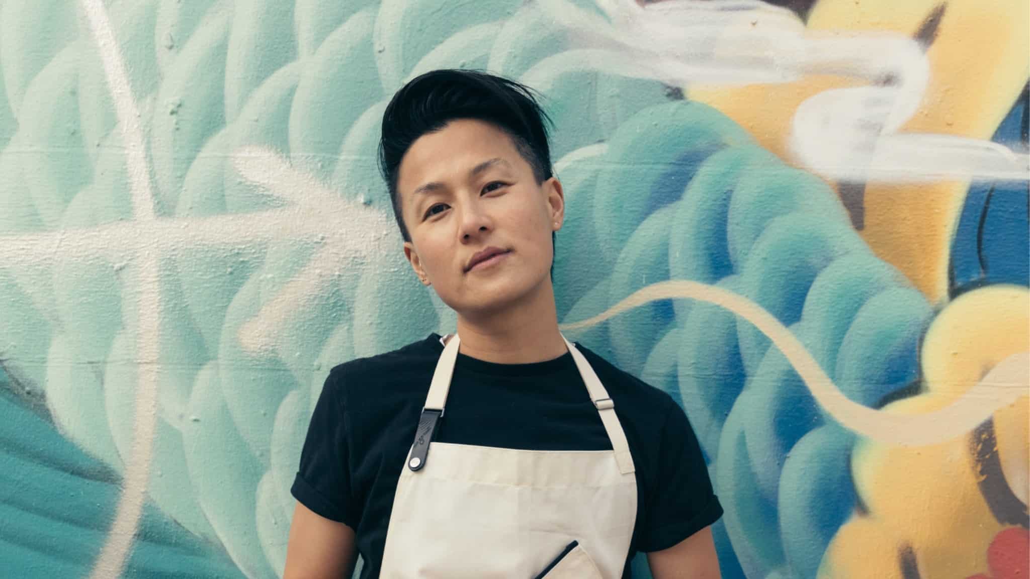 Person with short dark hair wearing a black t-shirt and a light apron stands in front of a colorful mural with swirling blue, white, and yellow patterns.