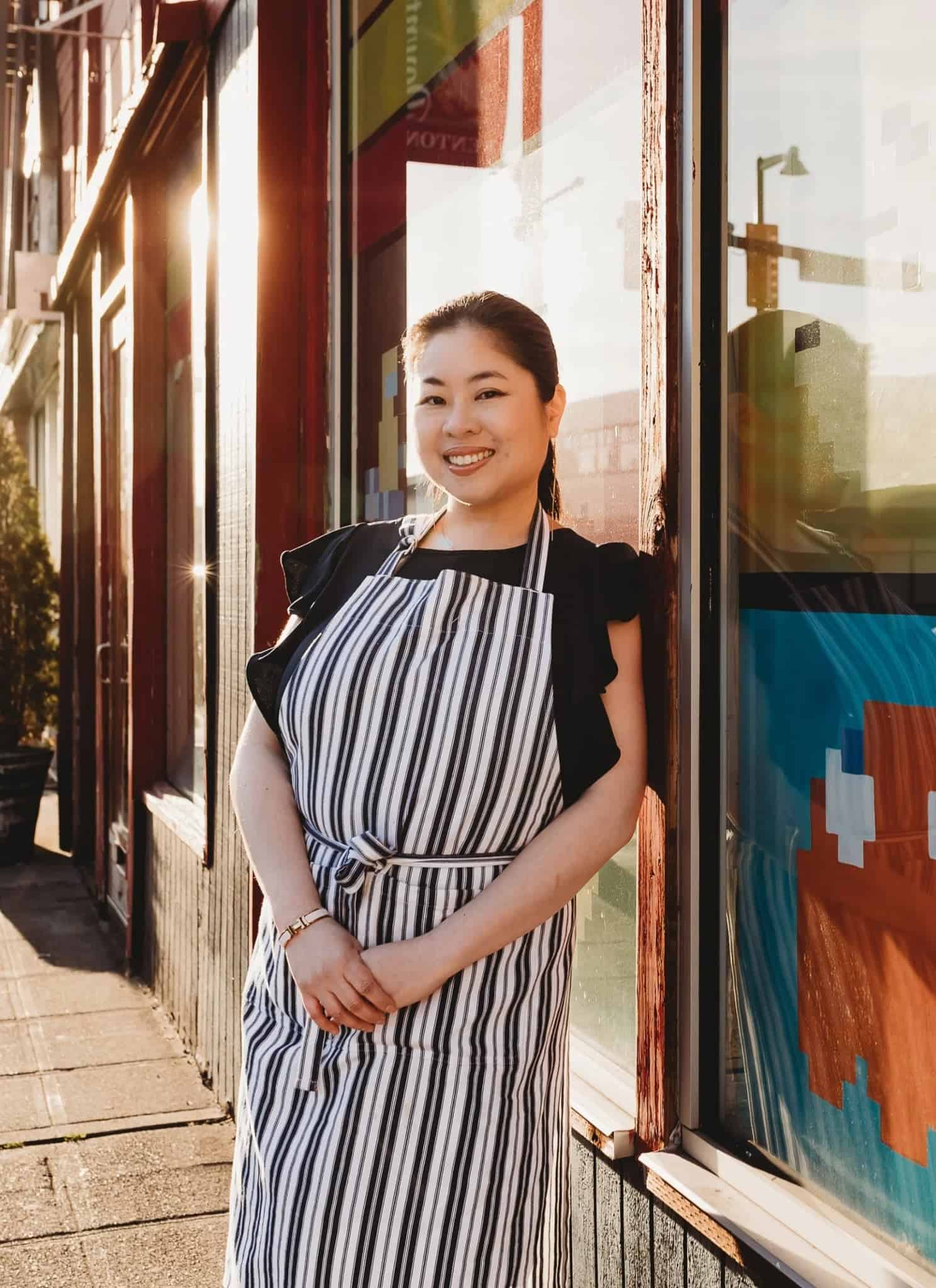 A woman in an apron smiles while leaning against a storefront window. Next to her is a book titled 108 Asian Cookies by Kat Lieu, with a green crinkle cookie on the cover.