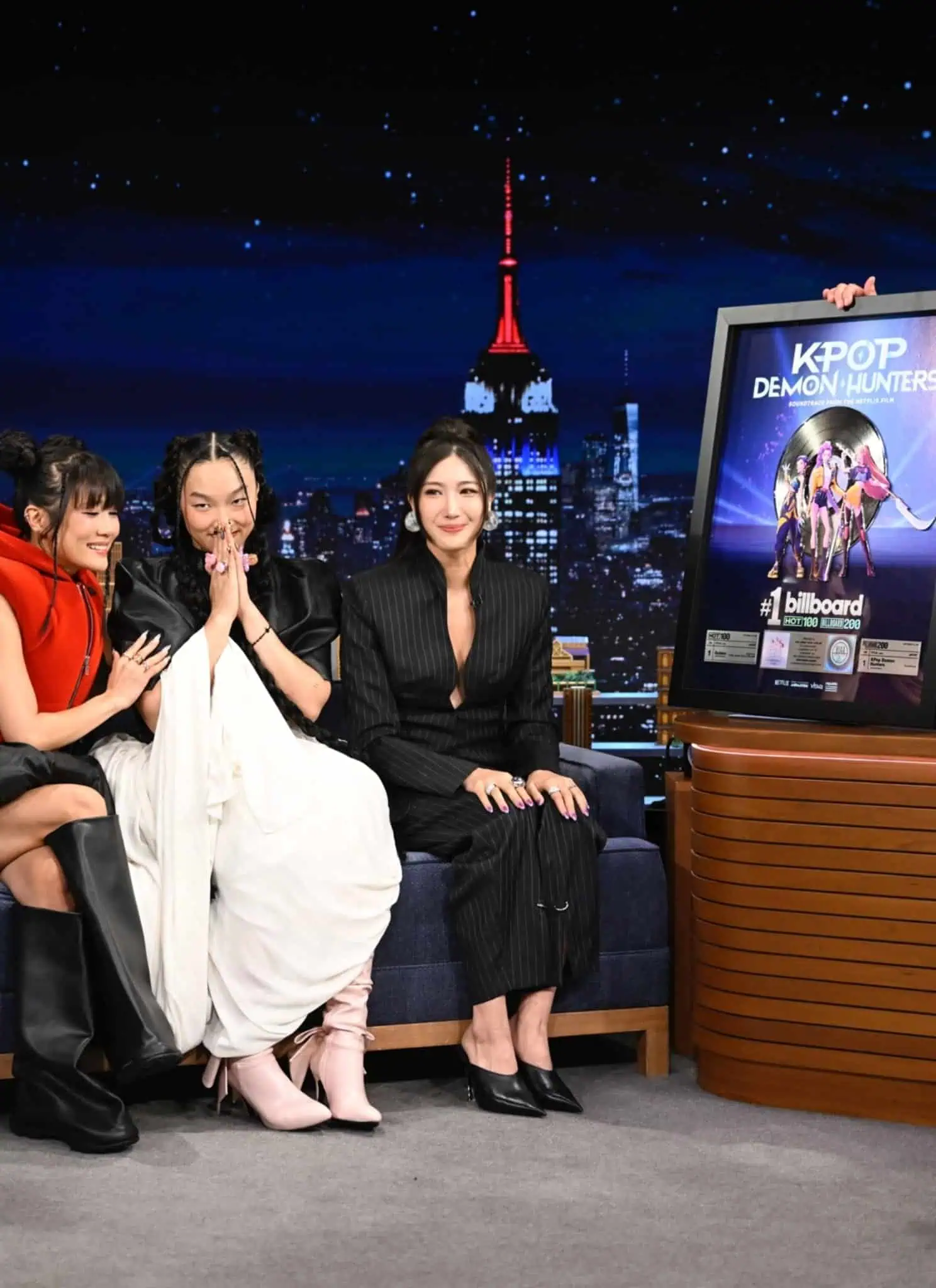 Three women sit on a talk show couch, smiling and posing, while a man in a suit stands beside them, holding a framed “K-Pop: Demon Hunters” poster. A cityscape is visible through the studio window behind them.