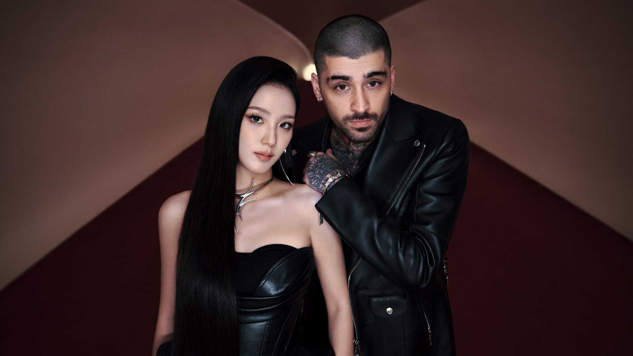 A woman with long black hair and a man with short hair and tattoos pose together in black leather outfits against a softly lit, geometric background.
