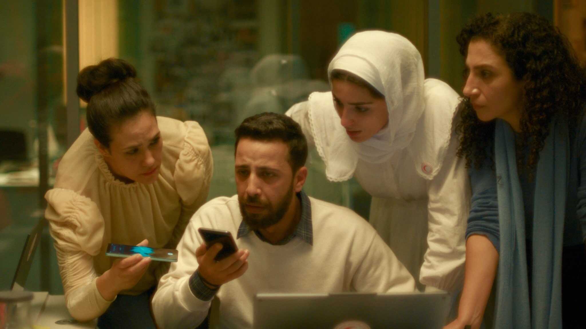 Four people gather around a table, focused on a smartphone held by a man sitting with a laptop. Three women, one in a white headscarf, lean in intently, suggesting teamwork or problem-solving.