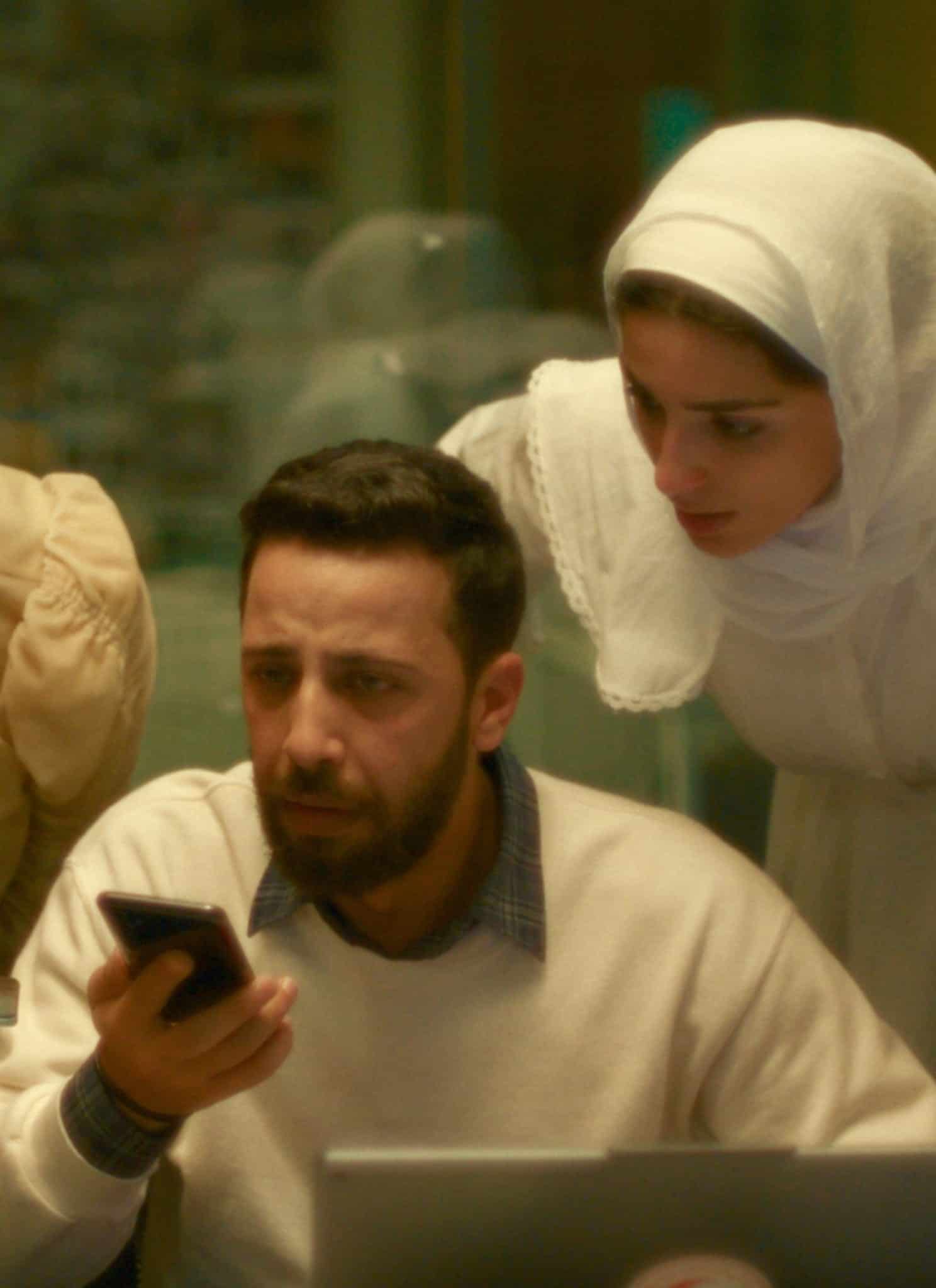 Four people gather around a table, focused on a smartphone held by a man sitting with a laptop. Three women, one in a white headscarf, lean in intently, suggesting teamwork or problem-solving.
