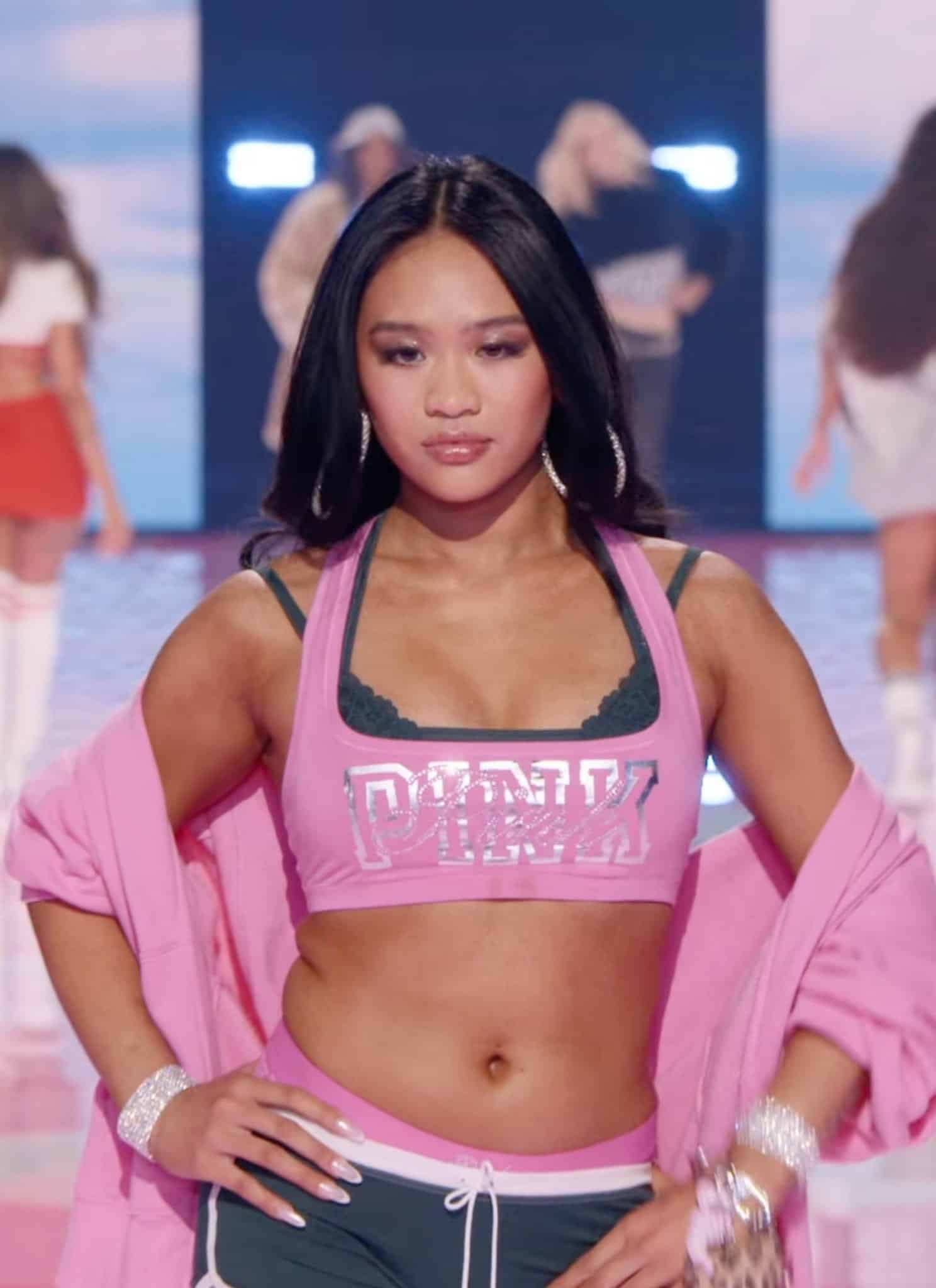 A model confidently walks a pink runway wearing a sporty pink crop top, black shorts, and a pink jacket while other models pose and dance in the background. The scene is vibrant and energetic.