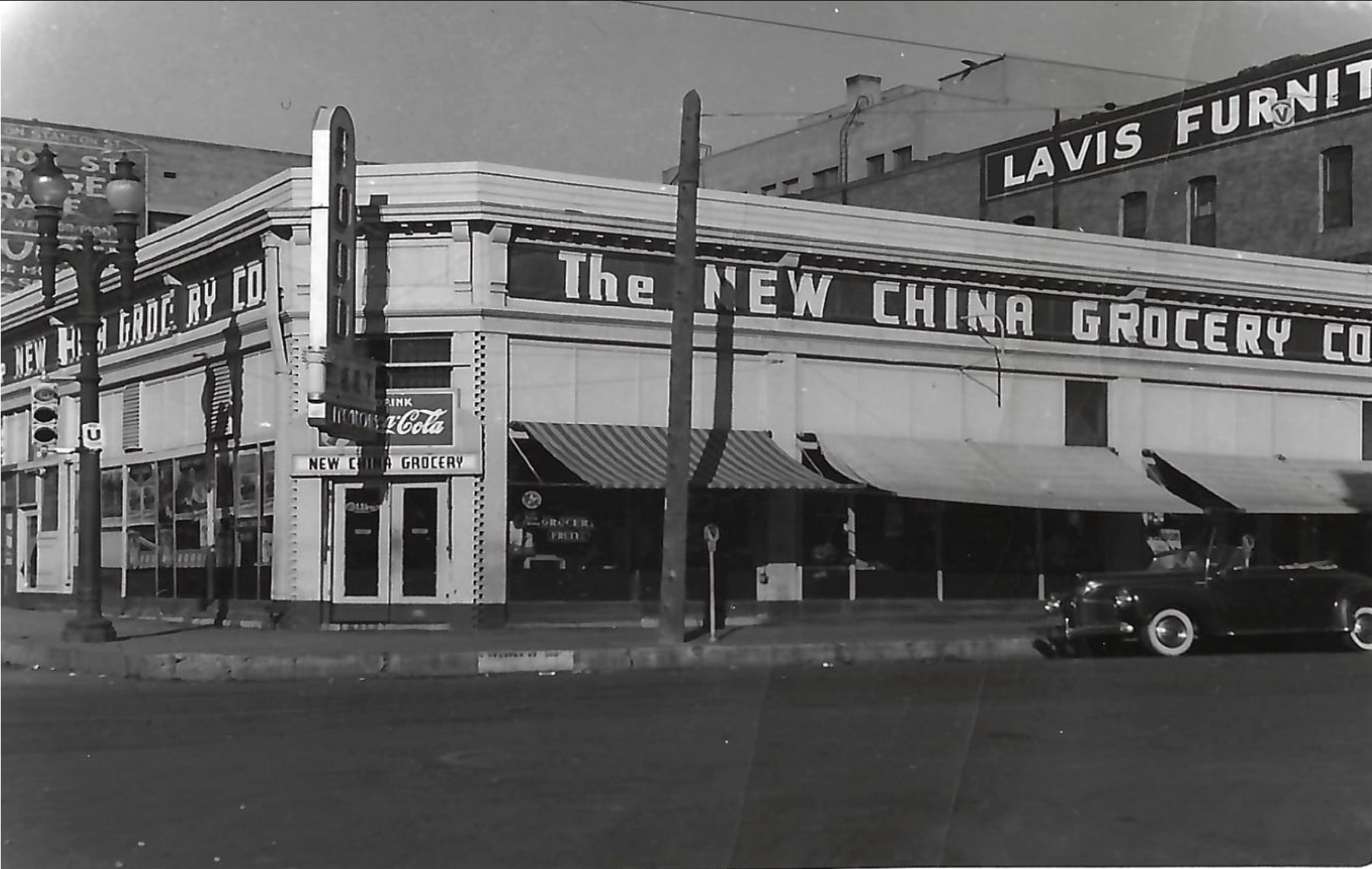Black-and-white photo of a corner building labeled The New China Grocery Co. A vintage car is parked at the curb, and nearby signs display “Lavis Furnit[ure]” and “Coca-Cola.” Awning shades cover the storefront windows.