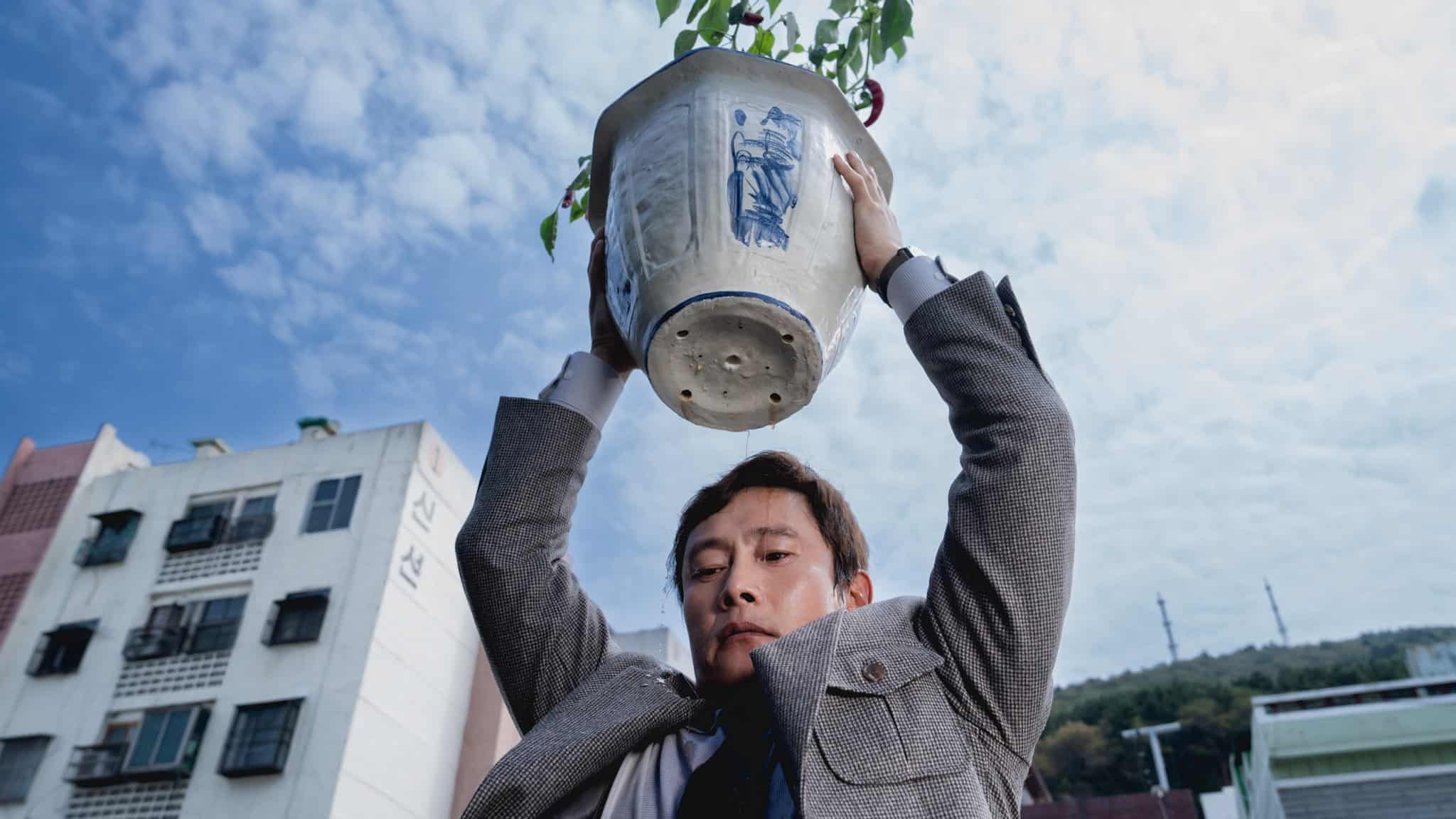 A man in a suit lifts a large ceramic flower pot above his head outdoors, with buildings and a partly cloudy sky in the background.