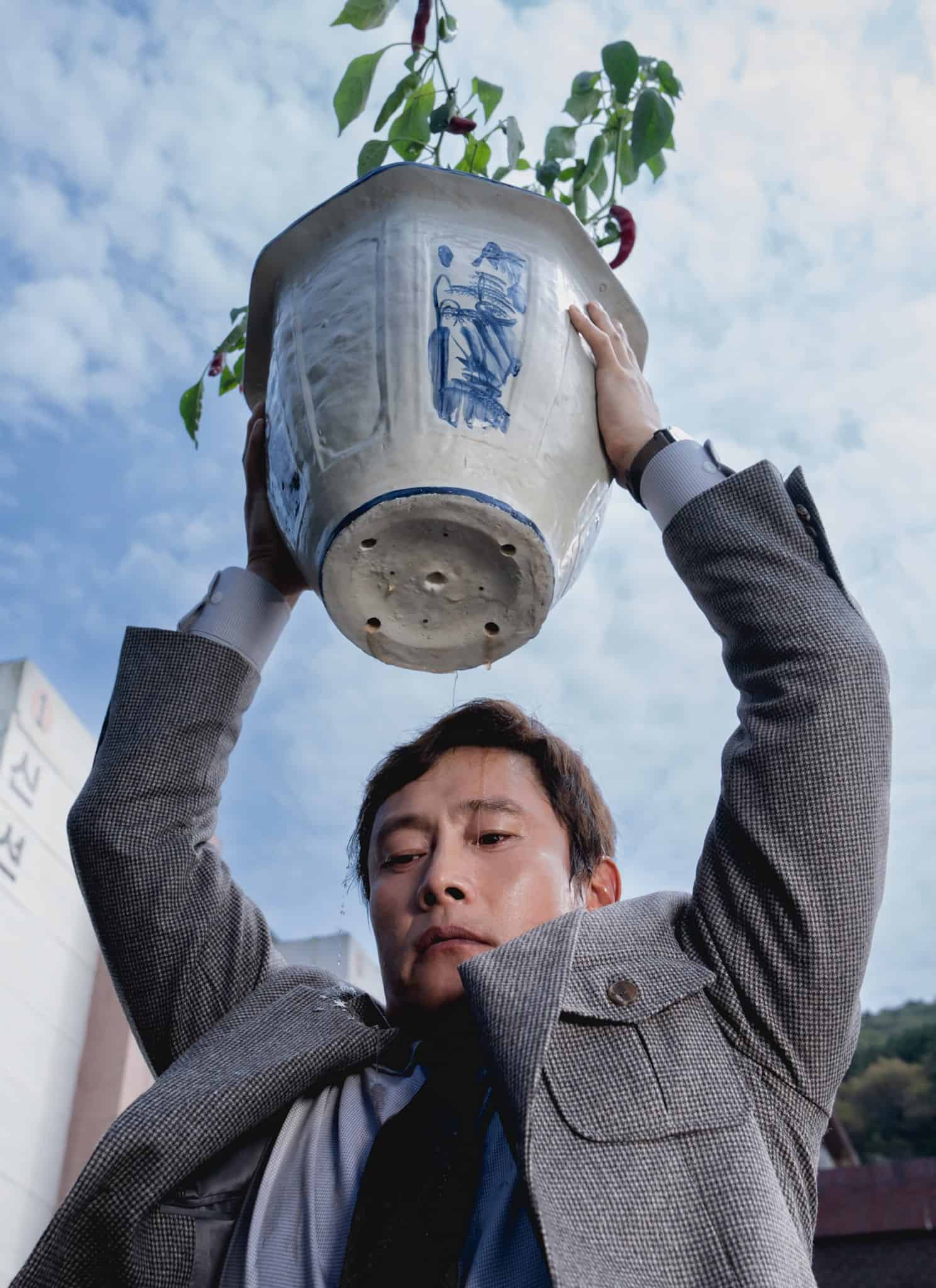 A man in a suit lifts a large ceramic flower pot above his head outdoors, with buildings and a partly cloudy sky in the background.
