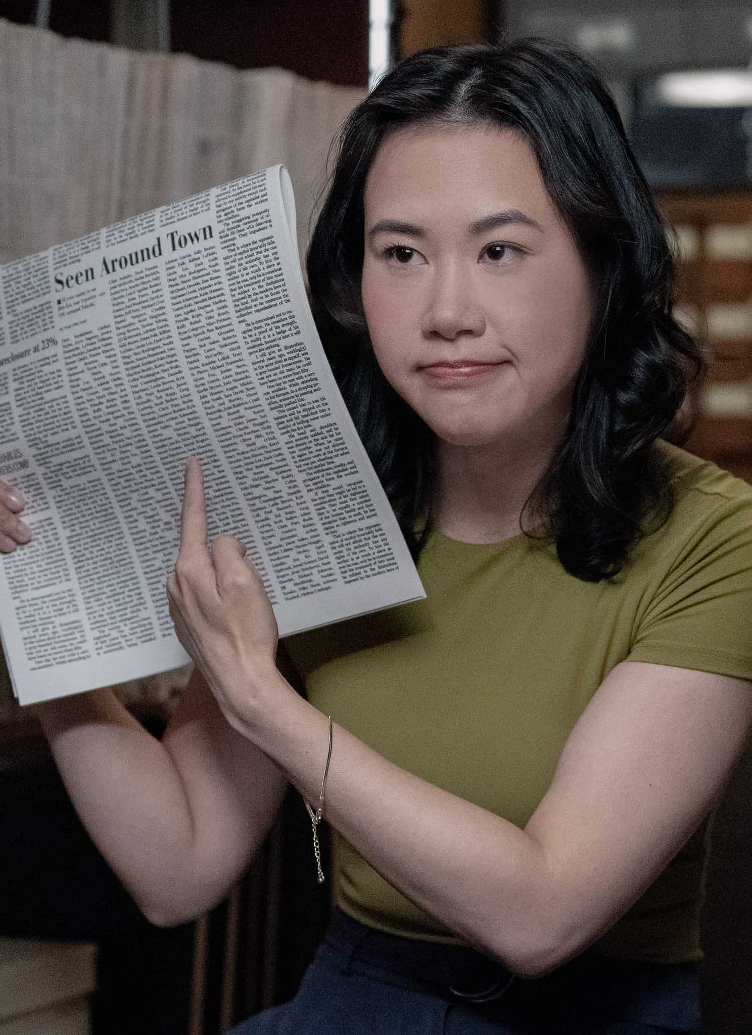 A woman with shoulder-length dark hair, wearing a green shirt, sits in an archive room and points to an article in a newspaper she is holding up. Shelves of files and drawers are visible in the background.