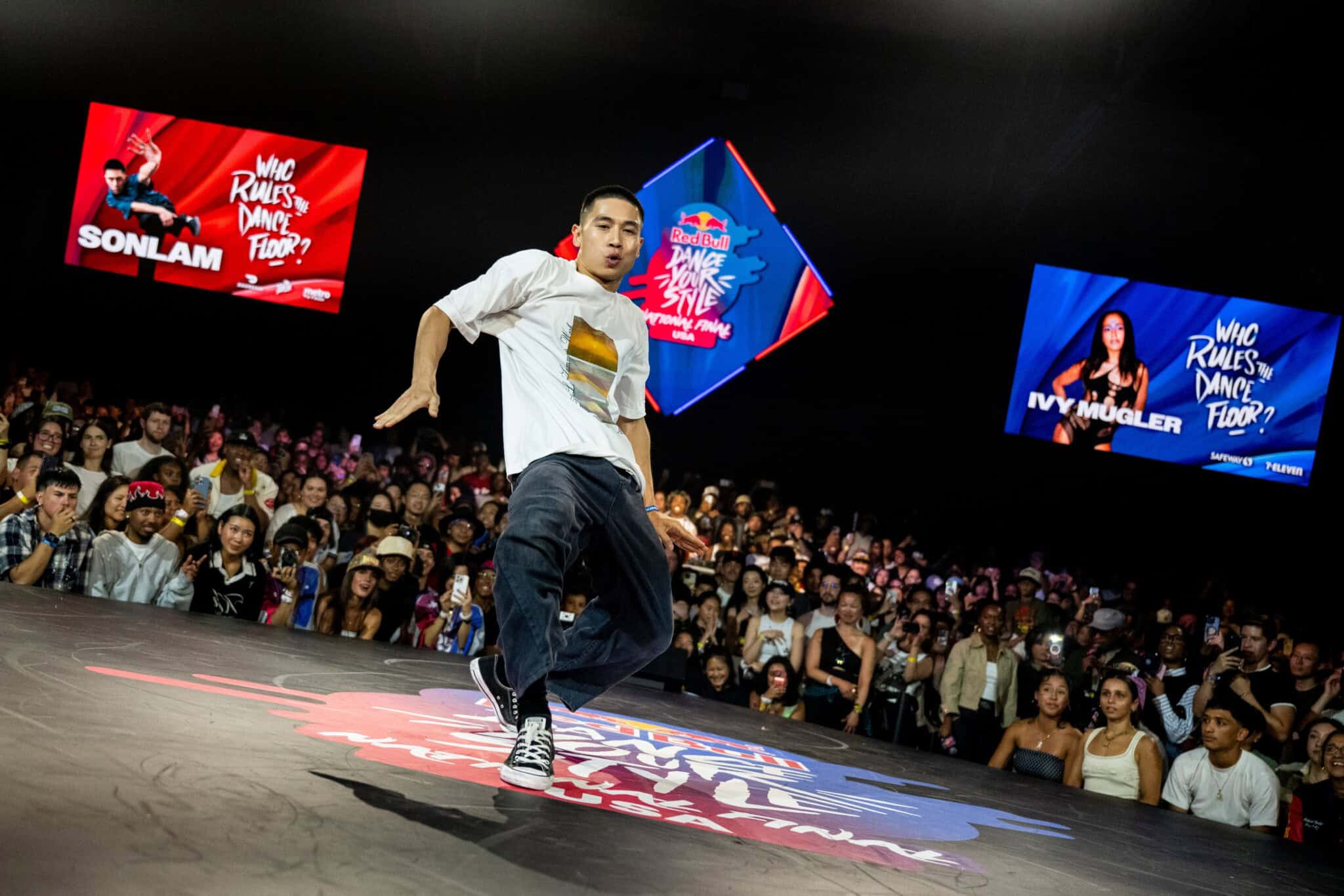 A street dancer performs energetically on stage at a competition, surrounded by a large, cheering audience. Large screens in the background display Who Rules the Dance Floor? and feature competitors’ names and images.
