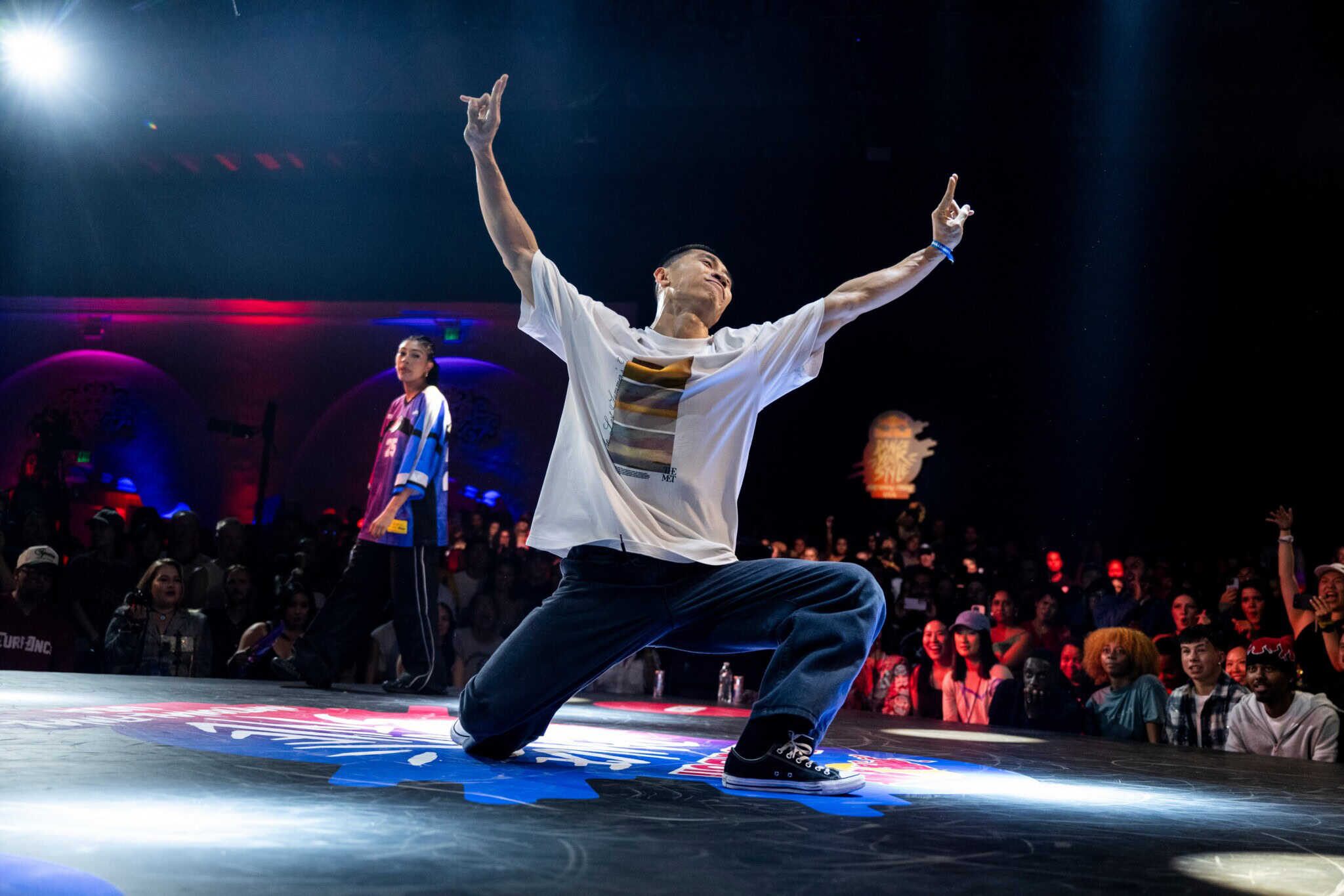 A breakdancer in a white shirt and blue jeans dramatically poses on one knee with arms raised on a stage, while a crowd watches and another dancer stands in the background under colorful lights.