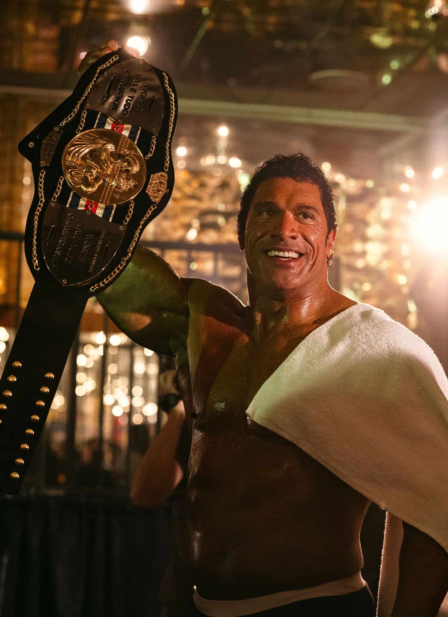 A smiling, sweaty man holds up a large wrestling championship belt with one arm, a white towel draped over his shoulder, as people applaud him in a brightly lit, celebratory setting.