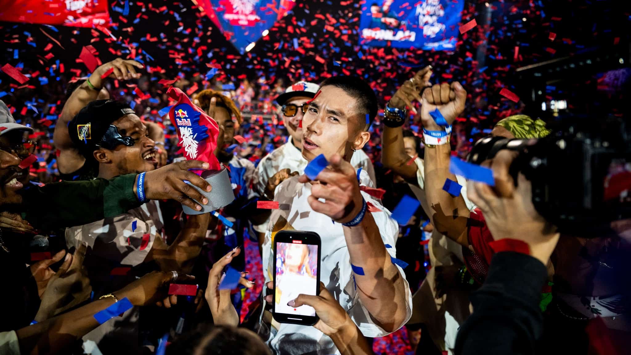 A man stands in the center surrounded by cheering people and blue and red confetti. Many are raising their hands and phones, capturing the moment as the man points forward, creating a lively, celebratory atmosphere.