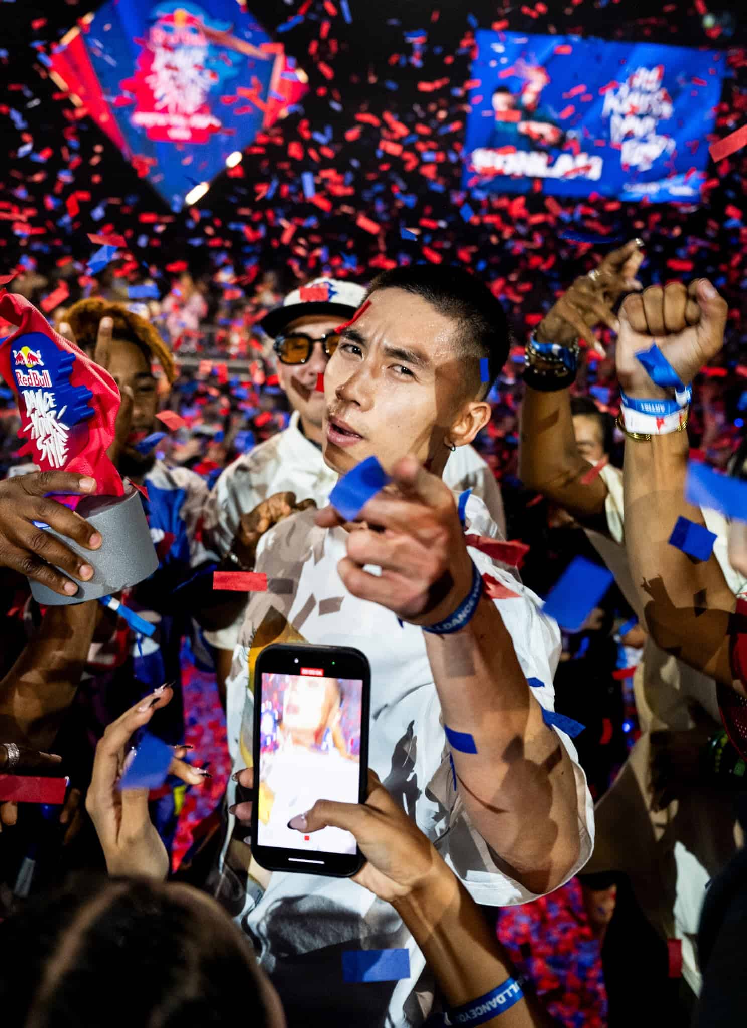 A man stands in the center surrounded by cheering people and blue and red confetti. Many are raising their hands and phones, capturing the moment as the man points forward, creating a lively, celebratory atmosphere.