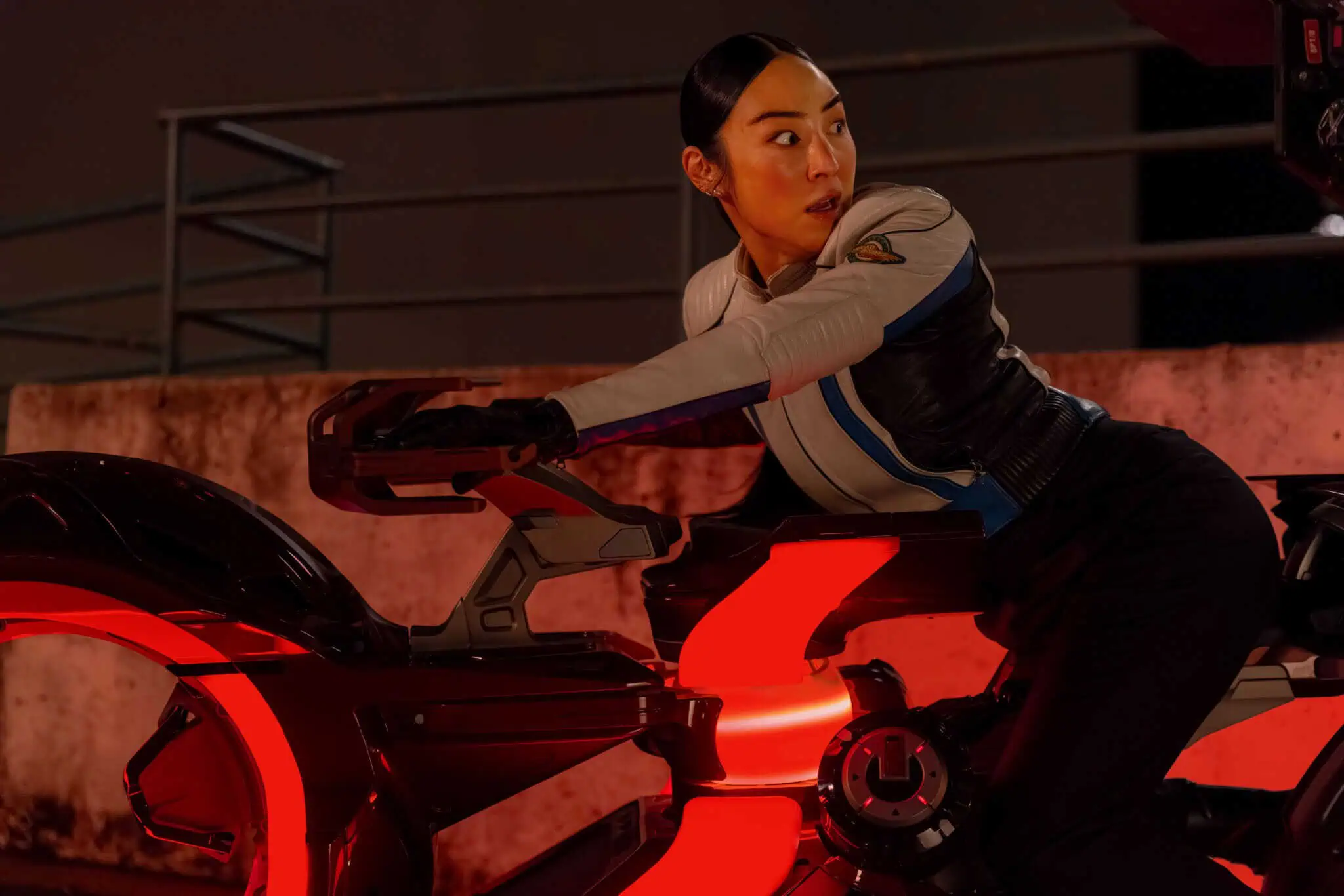A woman in a futuristic outfit sits on a glowing, high-tech motorcycle with red illuminated wheels, looking over her shoulder with a surprised expression. It appears to be nighttime and she is outdoors.