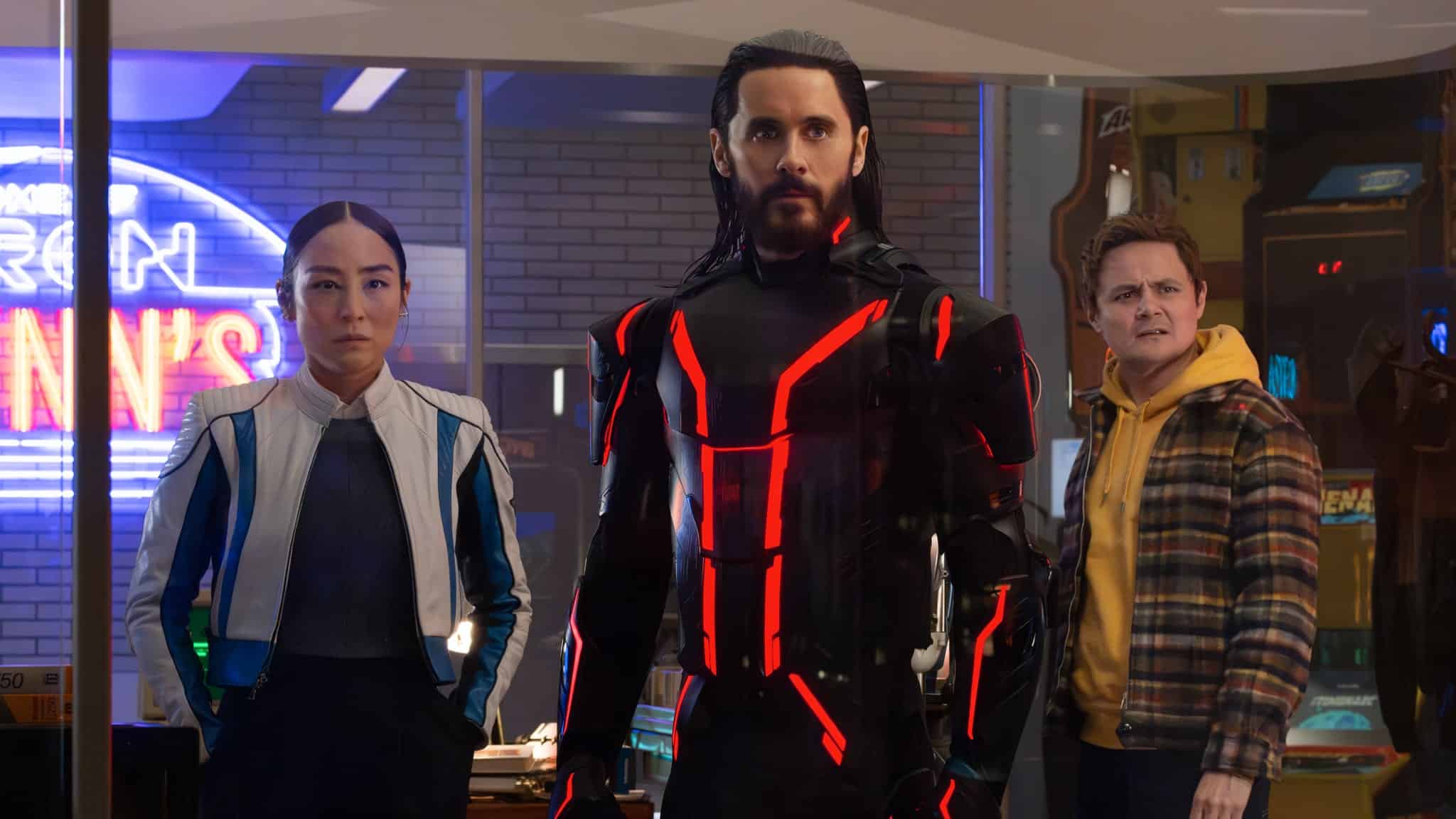 Three people stand indoors: a woman in a white and blue jacket, a man in a black suit with glowing red lines, and another man in a yellow plaid shirt, all with serious expressions, in a room with neon and arcade signs.