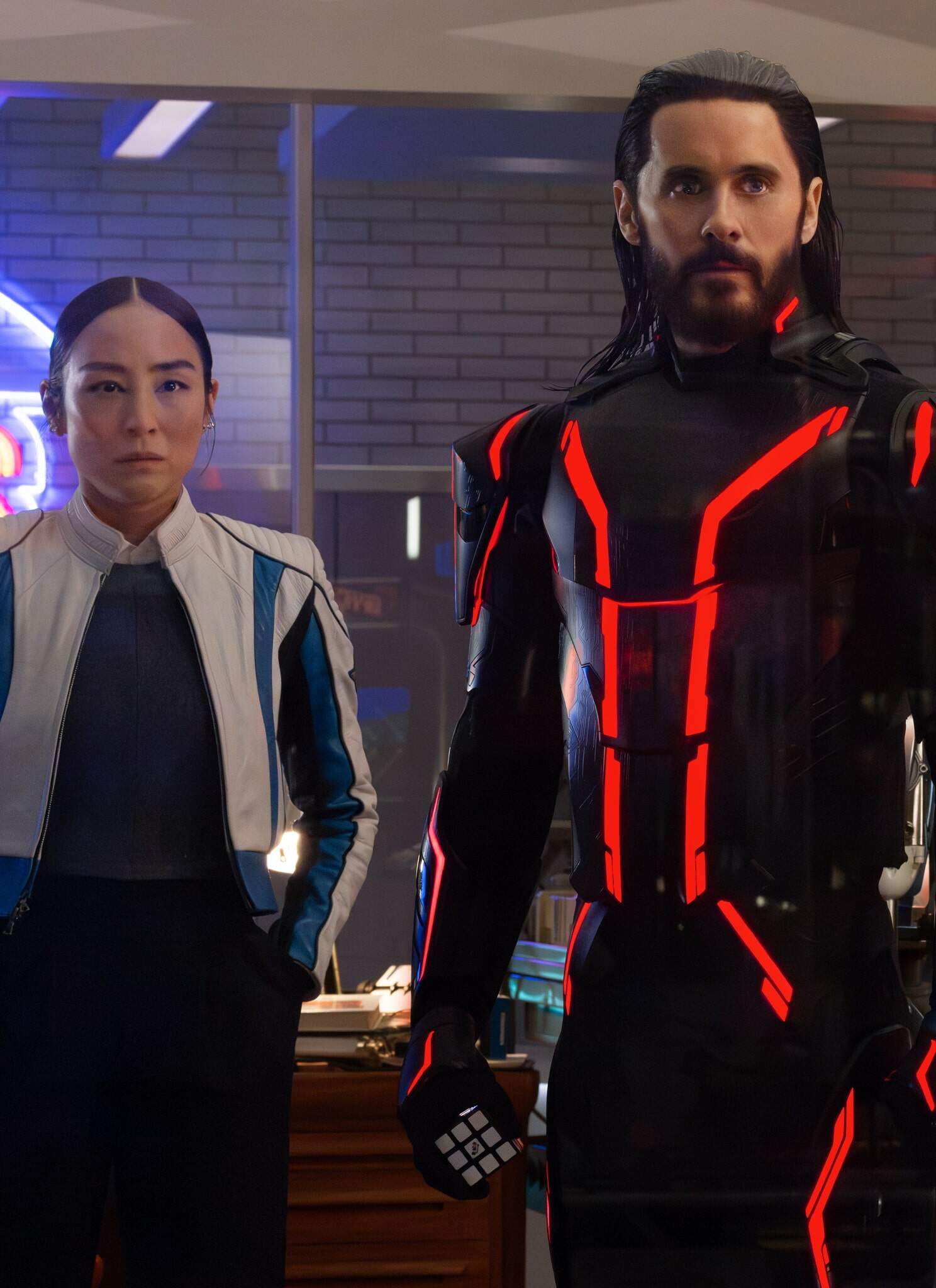 Three people stand indoors: a woman in a white and blue jacket, a man in a black suit with glowing red lines, and another man in a yellow plaid shirt, all with serious expressions, in a room with neon and arcade signs.