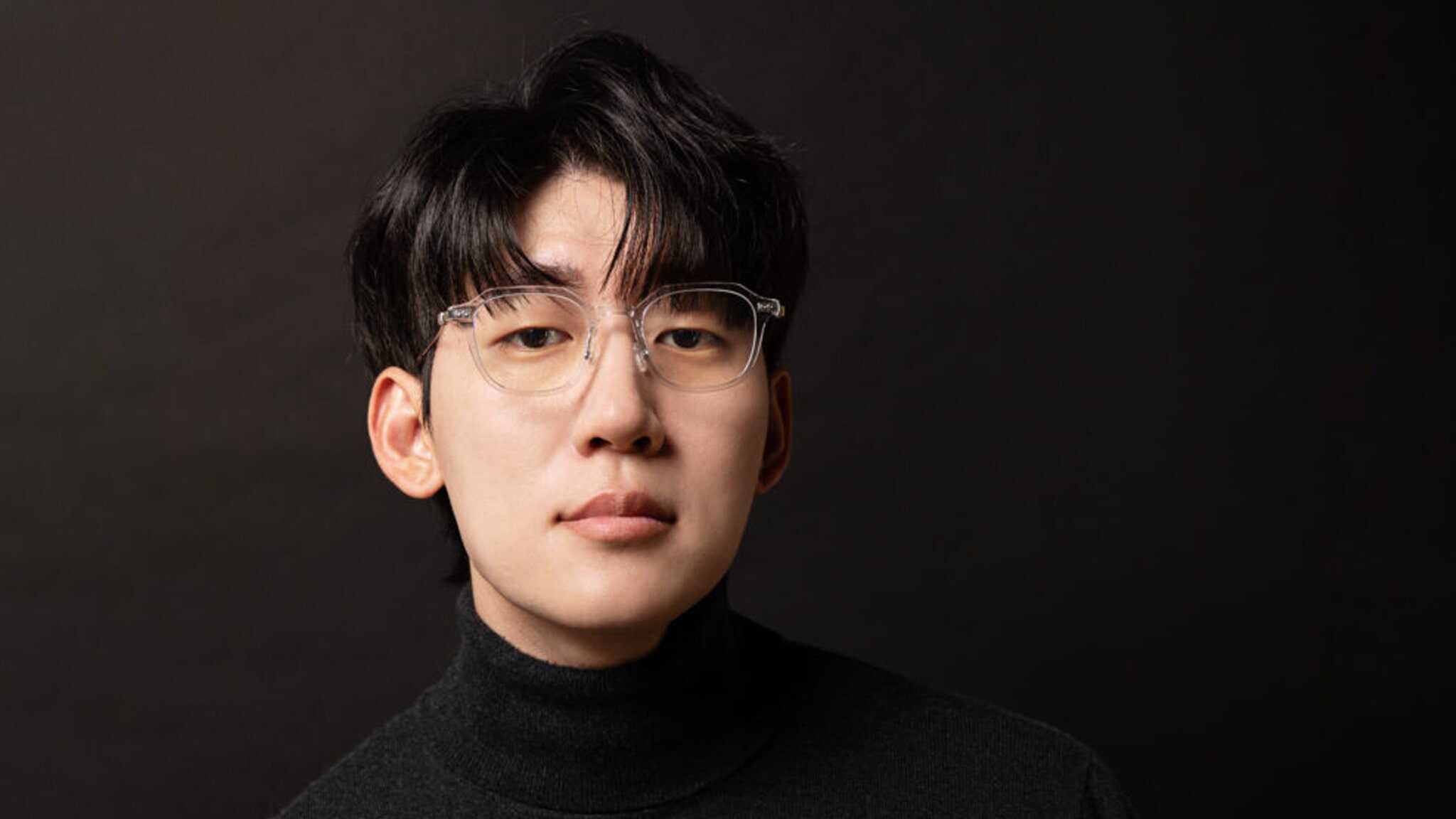 A young man with black hair and clear-framed glasses wearing a black turtleneck poses against a dark background, looking directly at the camera with a neutral expression.