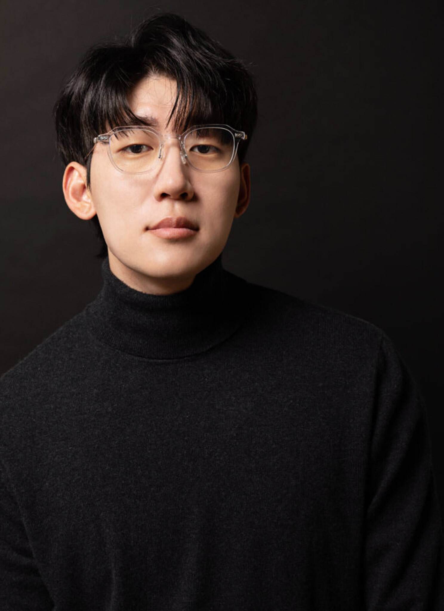 A young man with black hair and clear-framed glasses wearing a black turtleneck poses against a dark background, looking directly at the camera with a neutral expression.