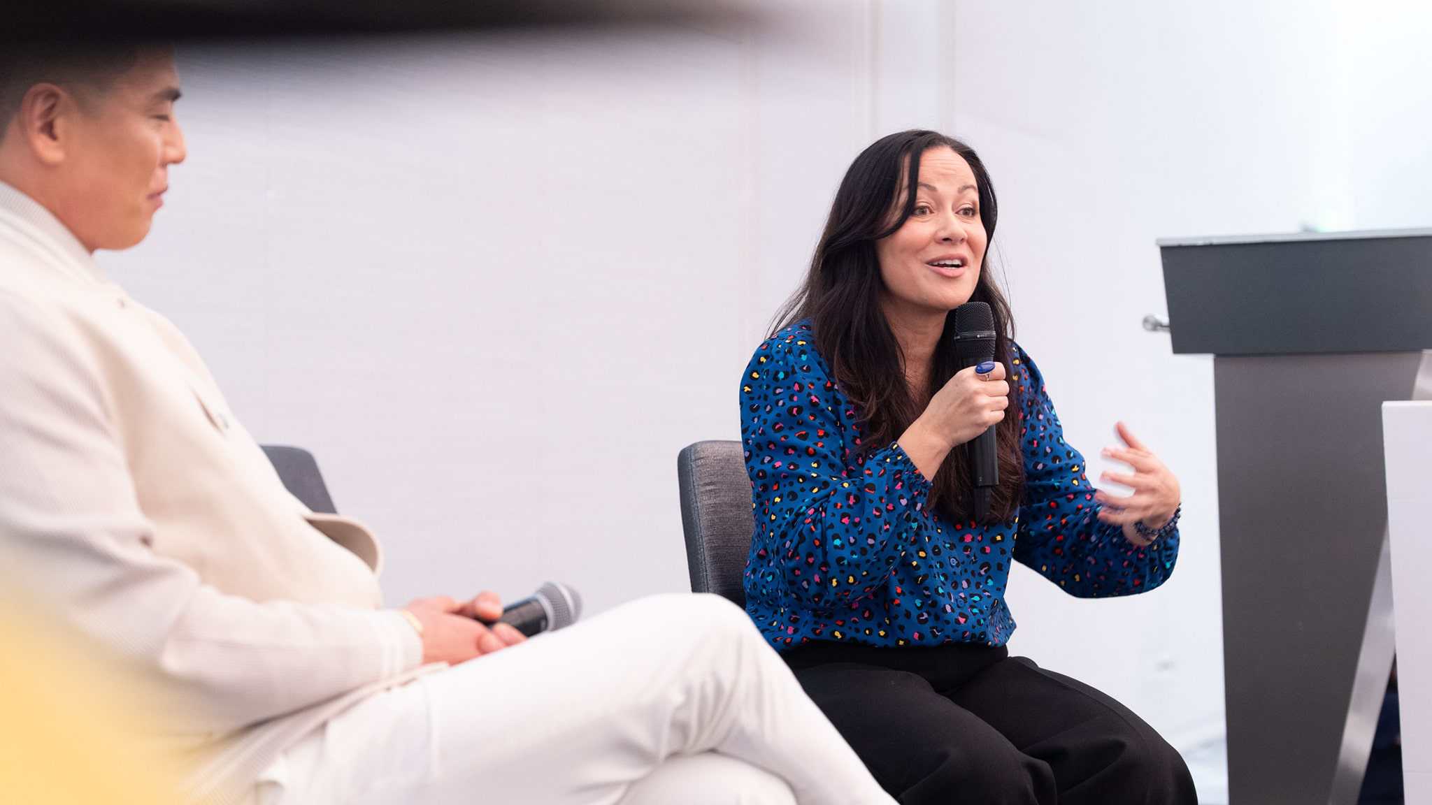A woman in a blue patterned shirt speaks into a microphone while gesturing with her hand. A man in a white outfit sits beside her, listening. They are seated on a stage in a brightly lit room.