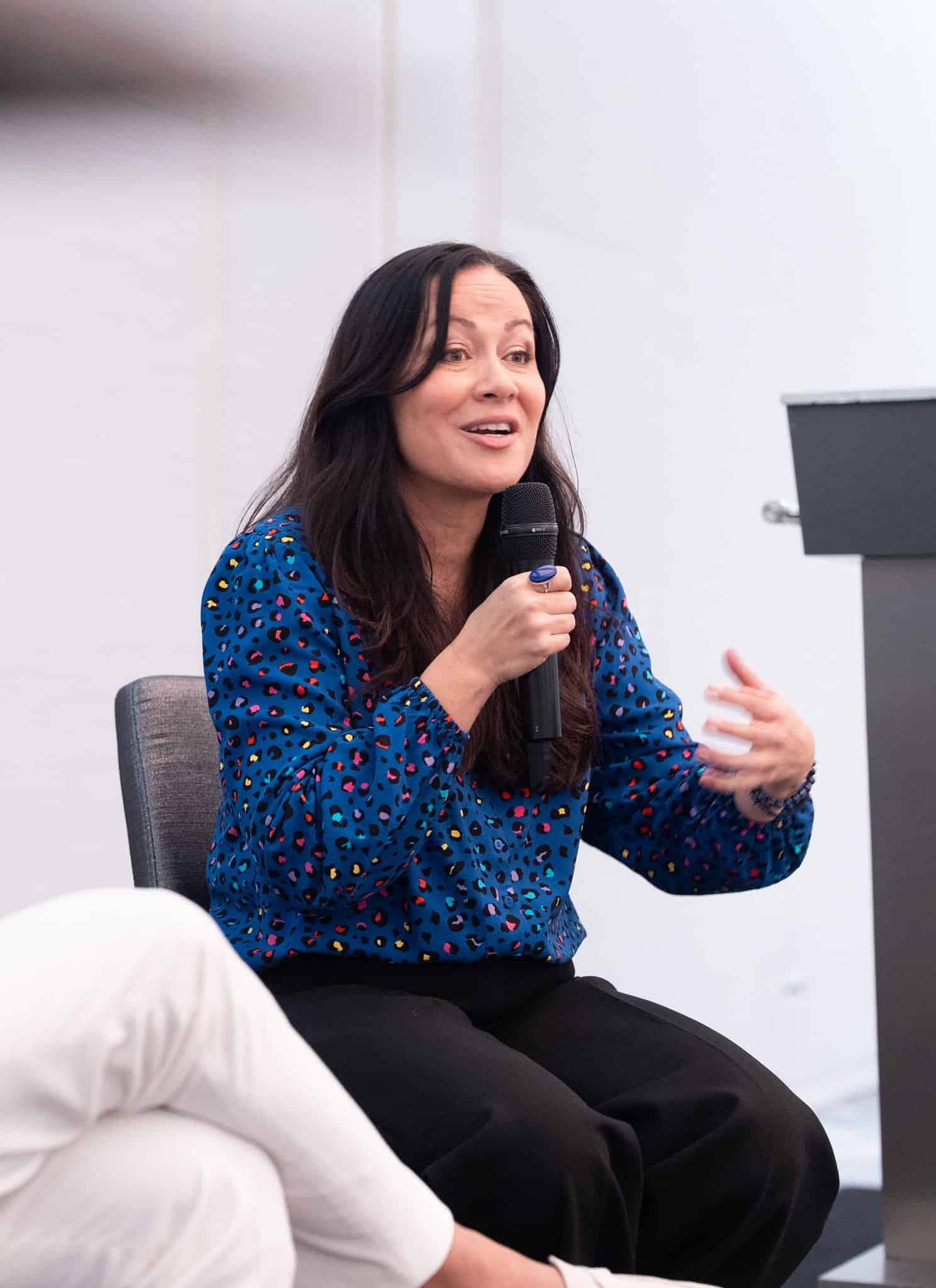 A woman in a blue patterned shirt speaks into a microphone while gesturing with her hand. A man in a white outfit sits beside her, listening. They are seated on a stage in a brightly lit room.