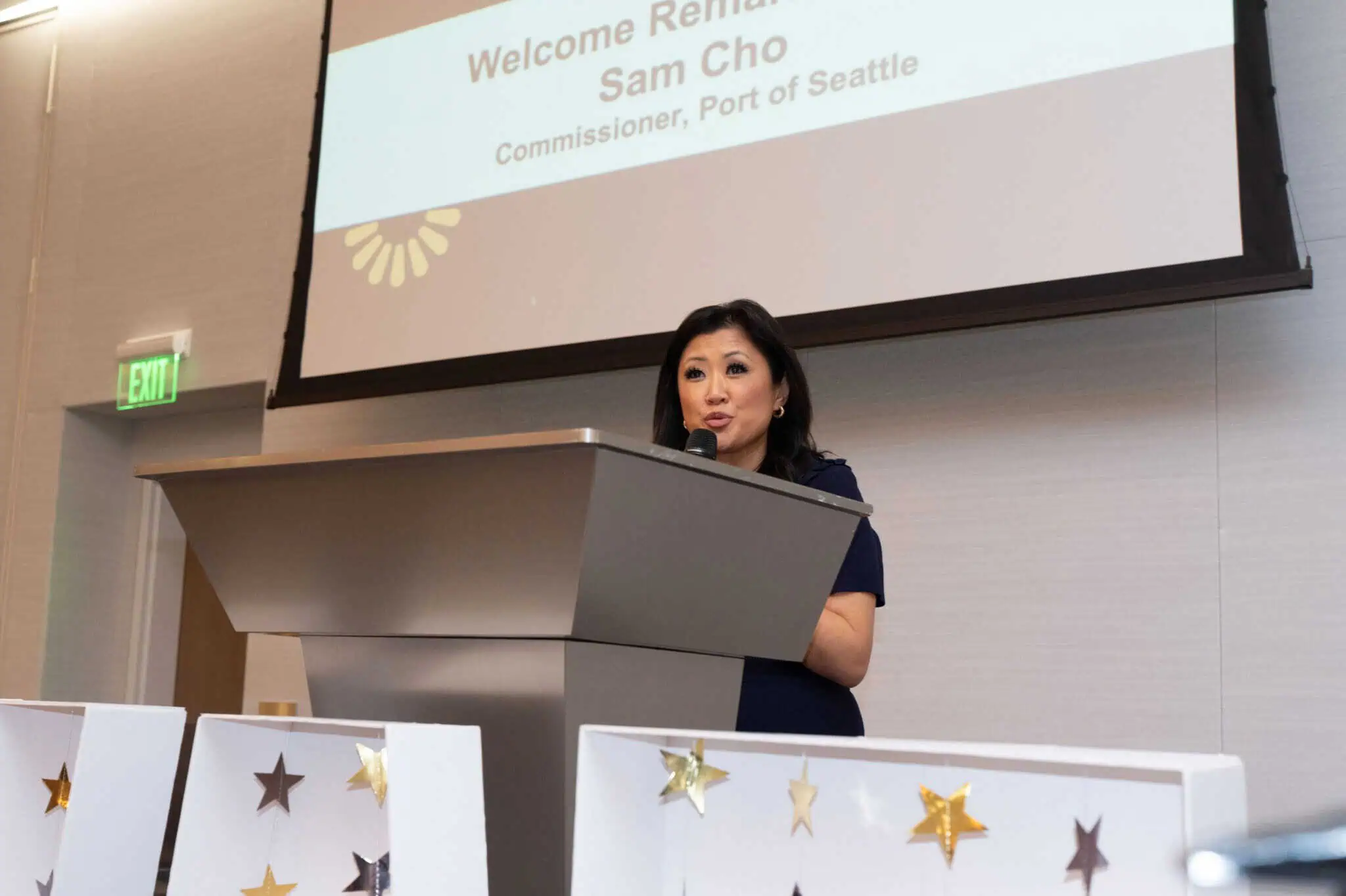 A woman stands at a podium speaking, with a projection screen behind her displaying the text Welcome Remarks Sam Cho, Commissioner, Port of Seattle. The podium is decorated with white panels and gold stars.