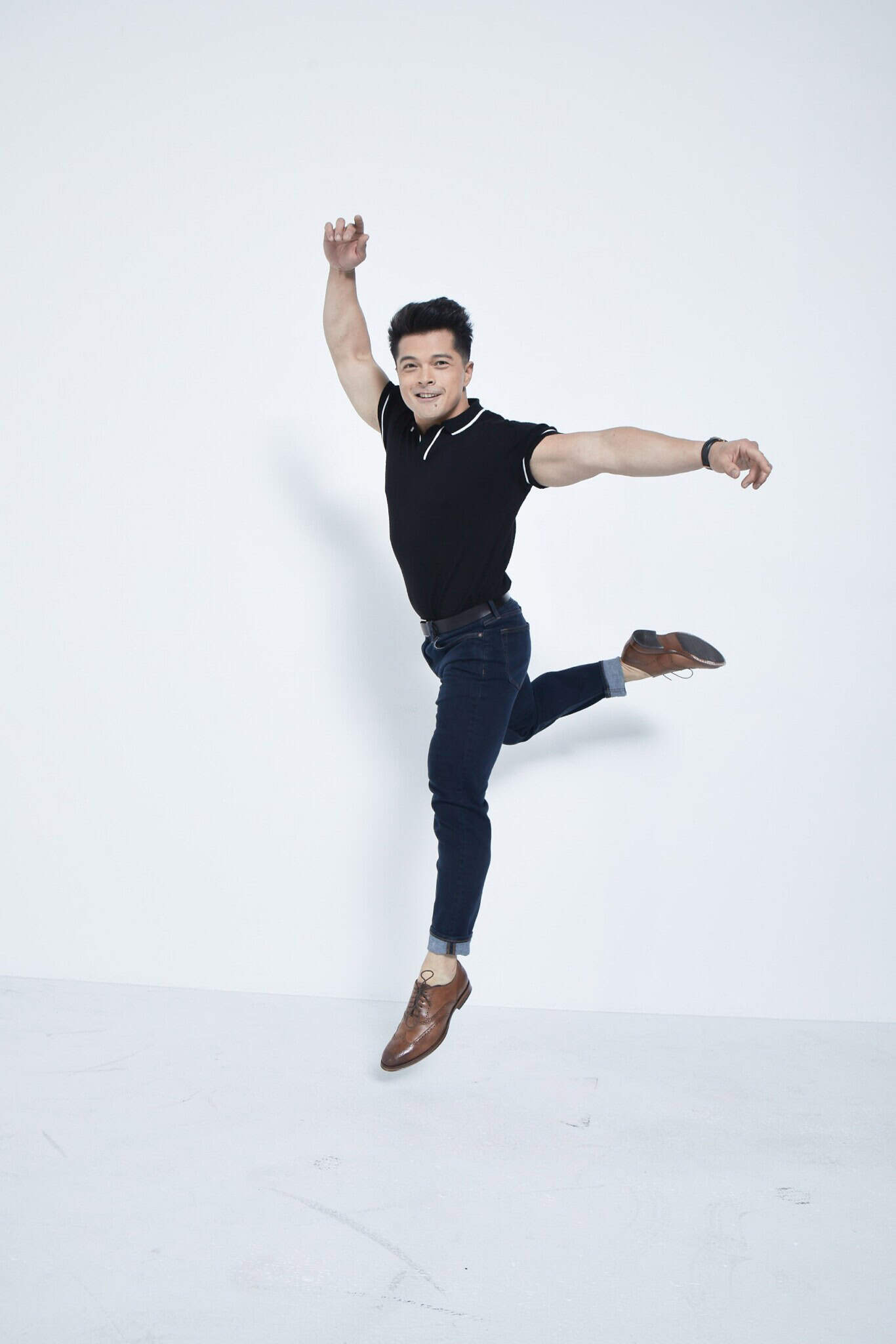 A man in a black polo shirt, dark jeans, and brown shoes poses mid-jump against a plain white background, with one arm raised and one leg bent behind him.