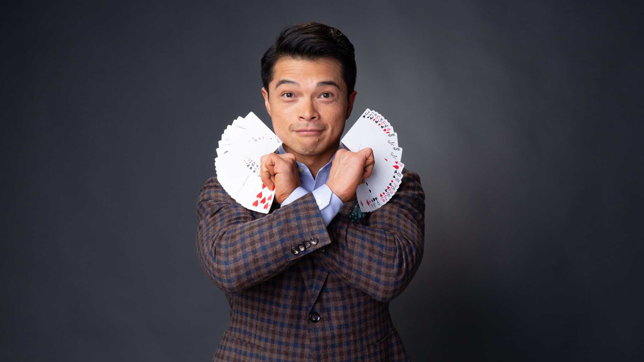 A man in a plaid suit jacket holds two fanned decks of playing cards, one in each hand, with his arms crossed in front of his chest against a plain dark background. He looks directly at the camera and smiles subtly.