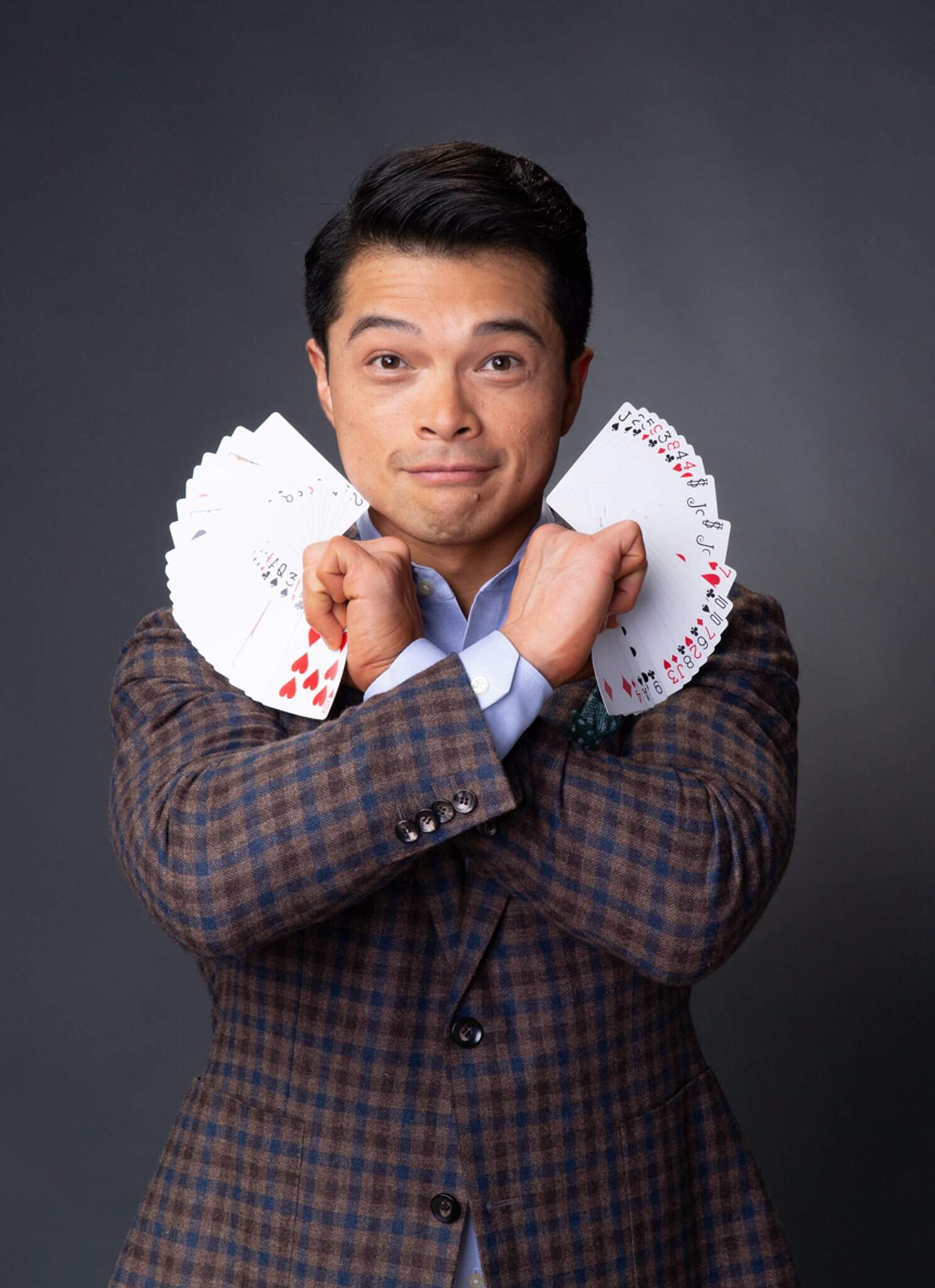 A man in a plaid suit jacket holds two fanned decks of playing cards, one in each hand, with his arms crossed in front of his chest against a plain dark background. He looks directly at the camera and smiles subtly.