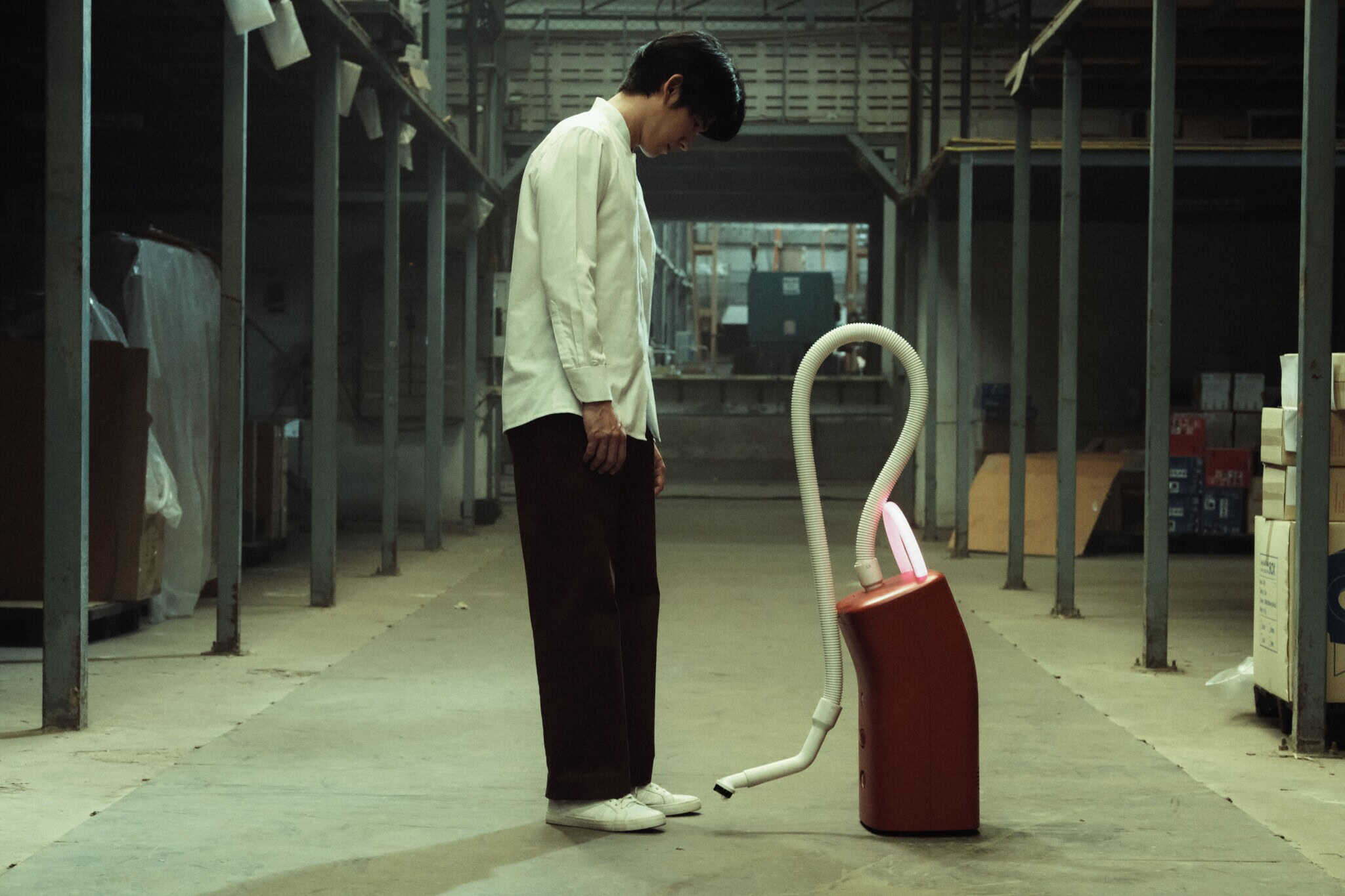 A person in a white shirt and dark pants stands in an industrial warehouse, looking down at a red vacuum cleaner with a raised hose pointed toward their foot.