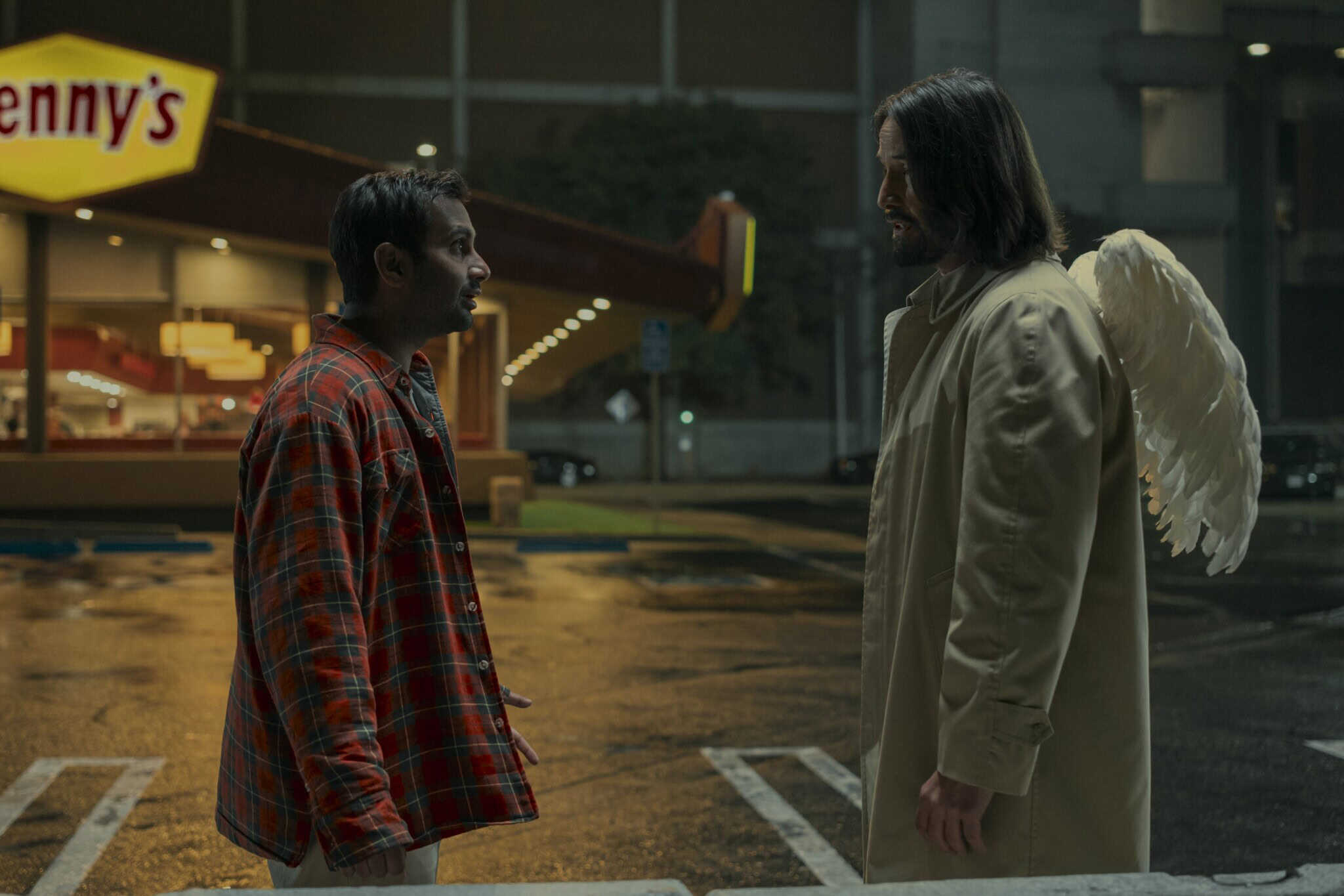 A man in a red plaid shirt faces another man with long hair, wearing a beige coat and white angel wings, in a wet, empty parking lot at night outside a Dennys restaurant.