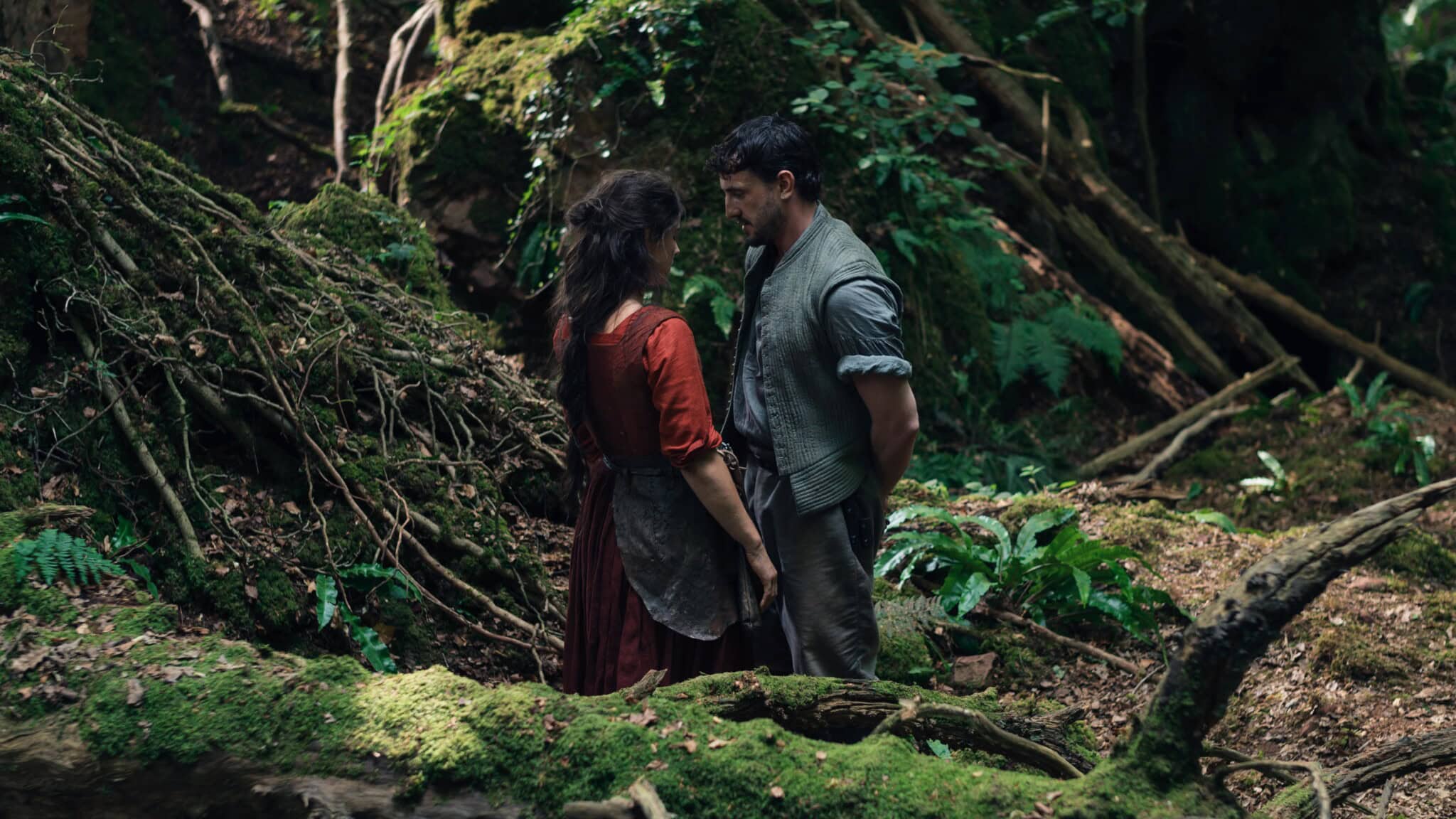 A man and woman stand close together facing each other in a lush, green forest, surrounded by moss-covered logs and dense foliage. The scene looks intimate and dramatic.