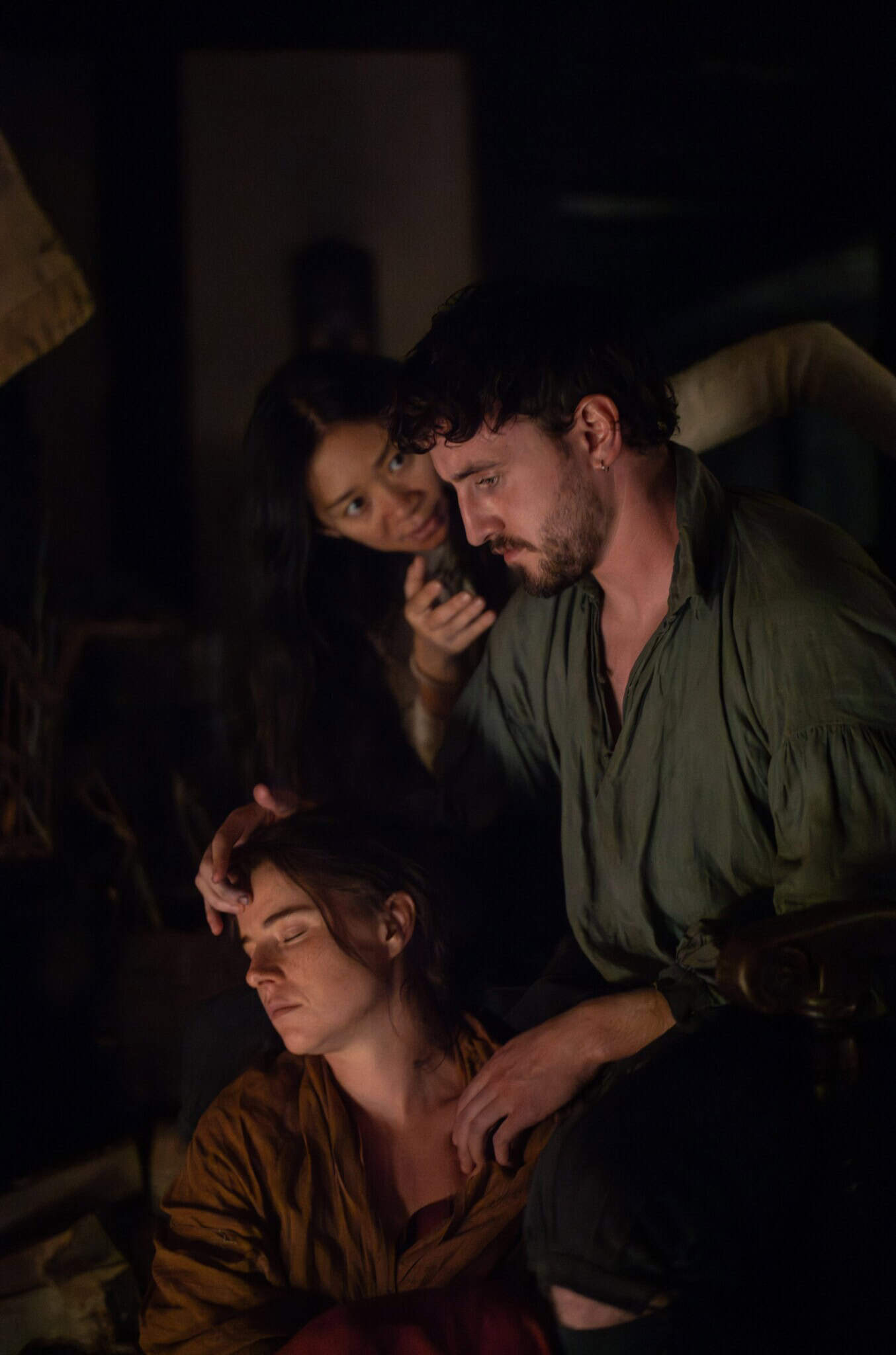 A man in a green shirt sits next to a woman resting her head on his knee, his hand gently on her head. Another woman stands behind them, appearing to comfort or speak to him in a dimly lit, intimate setting.