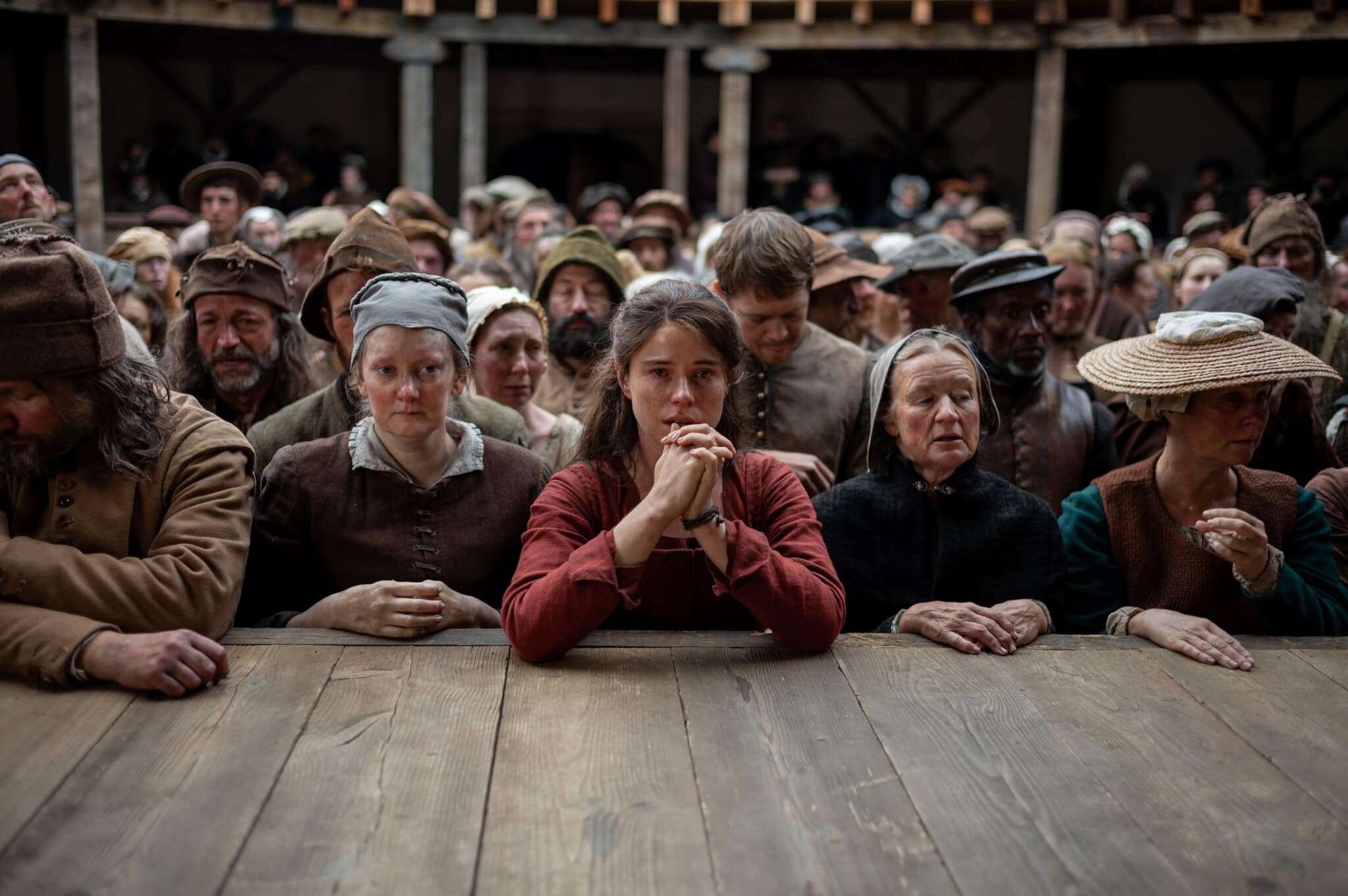 A crowd of people in historical clothing stand closely together behind a wooden railing, with a young woman in a red dress at the center, clasping her hands and looking forward intently.