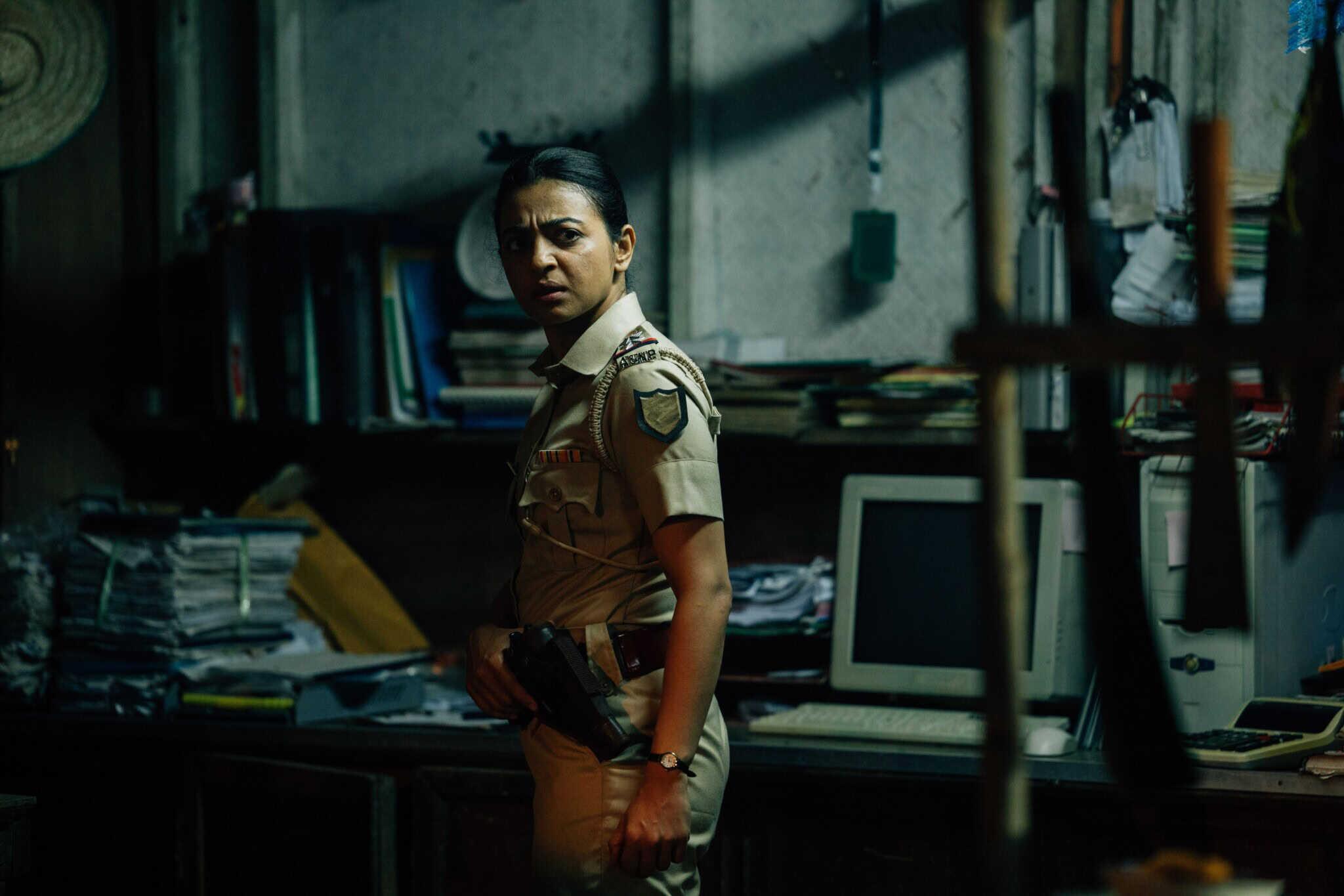 A woman in a police uniform stands in a dimly lit, cluttered office, holding a gun with a serious expression. There are stacks of papers, books, and an old computer on the desk behind her.