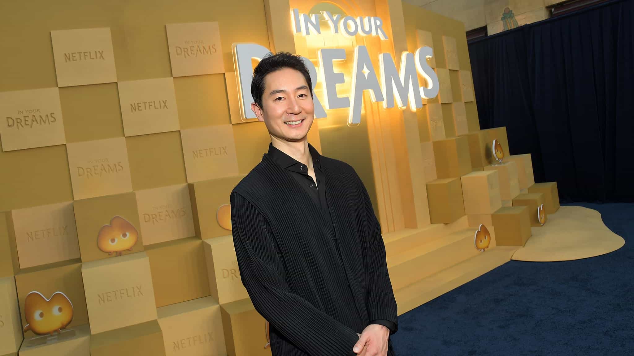 A man in a black outfit smiles while standing in front of a yellow backdrop with the words IN YOUR DREAMS and stacked boxes featuring Netflix and smiling blob logos.