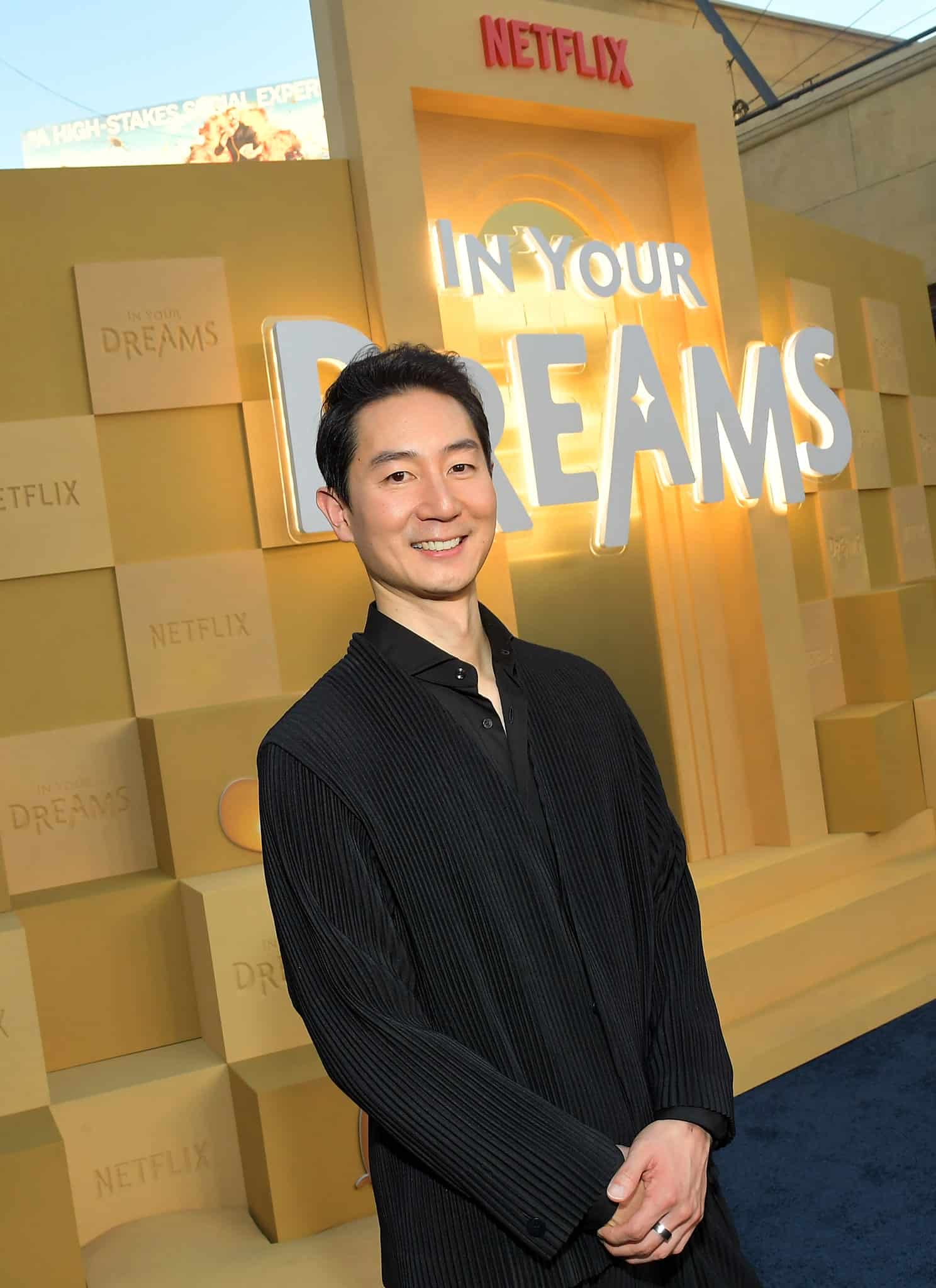 A man in a black outfit smiles while standing in front of a yellow backdrop with the words IN YOUR DREAMS and stacked boxes featuring Netflix and smiling blob logos.
