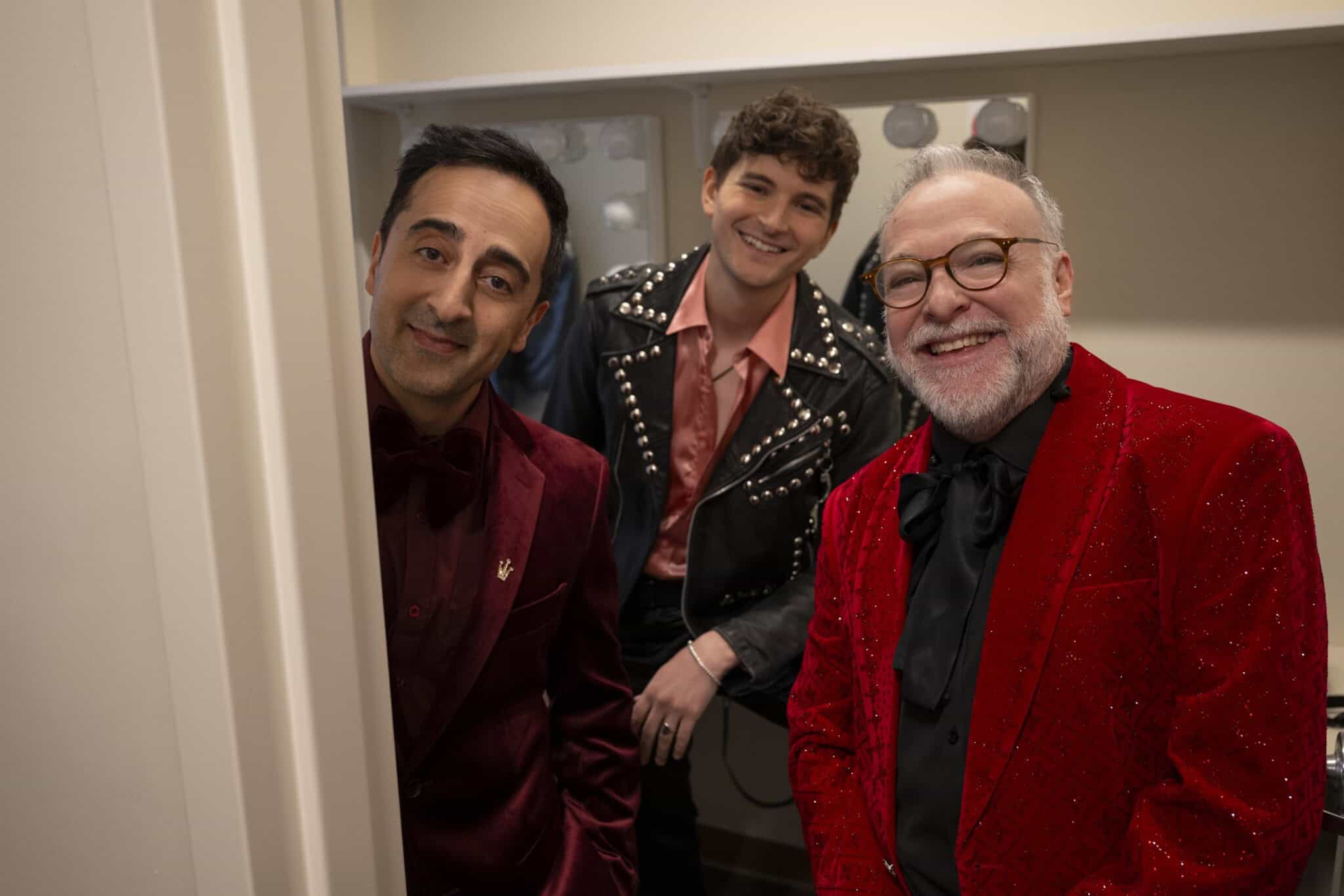 Three men smiling in a backstage dressing room. Two wear red velvet jackets, one with glasses, and the third wears a black studded leather jacket. They stand close together, appearing happy and relaxed.