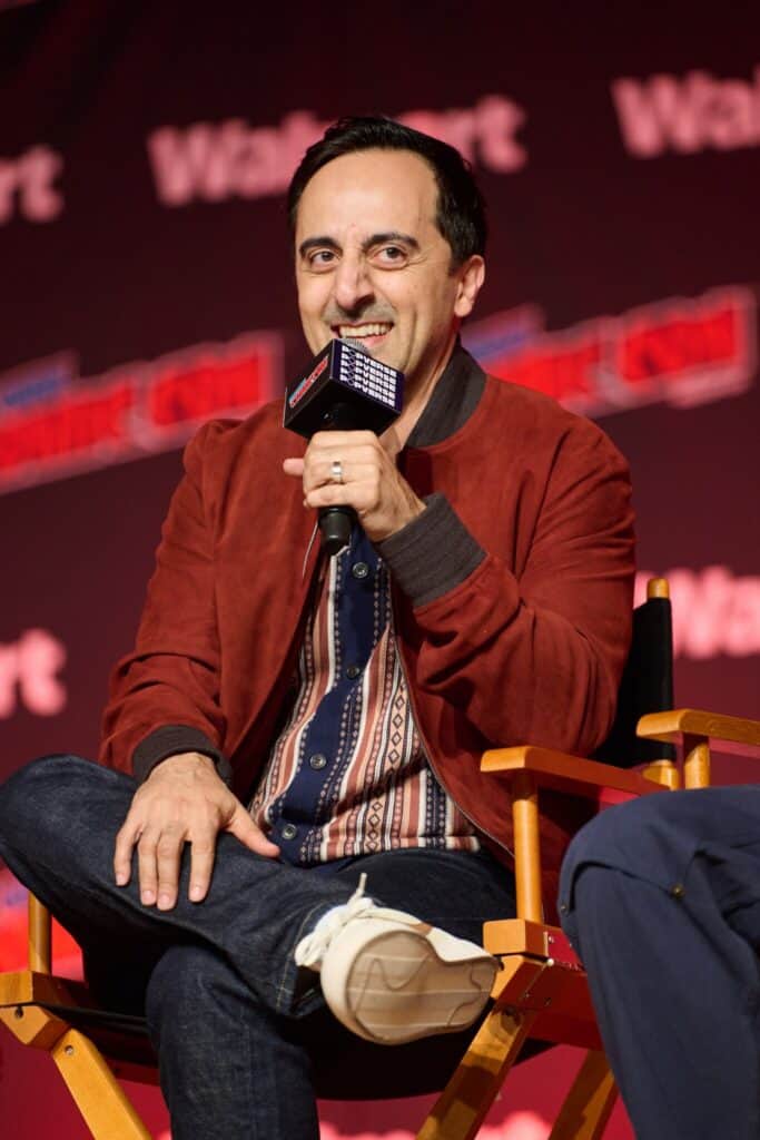 A man in a red jacket and patterned shirt sits on a directors chair, smiling and holding a microphone at a panel event with a red and black background displaying Walmart branding.