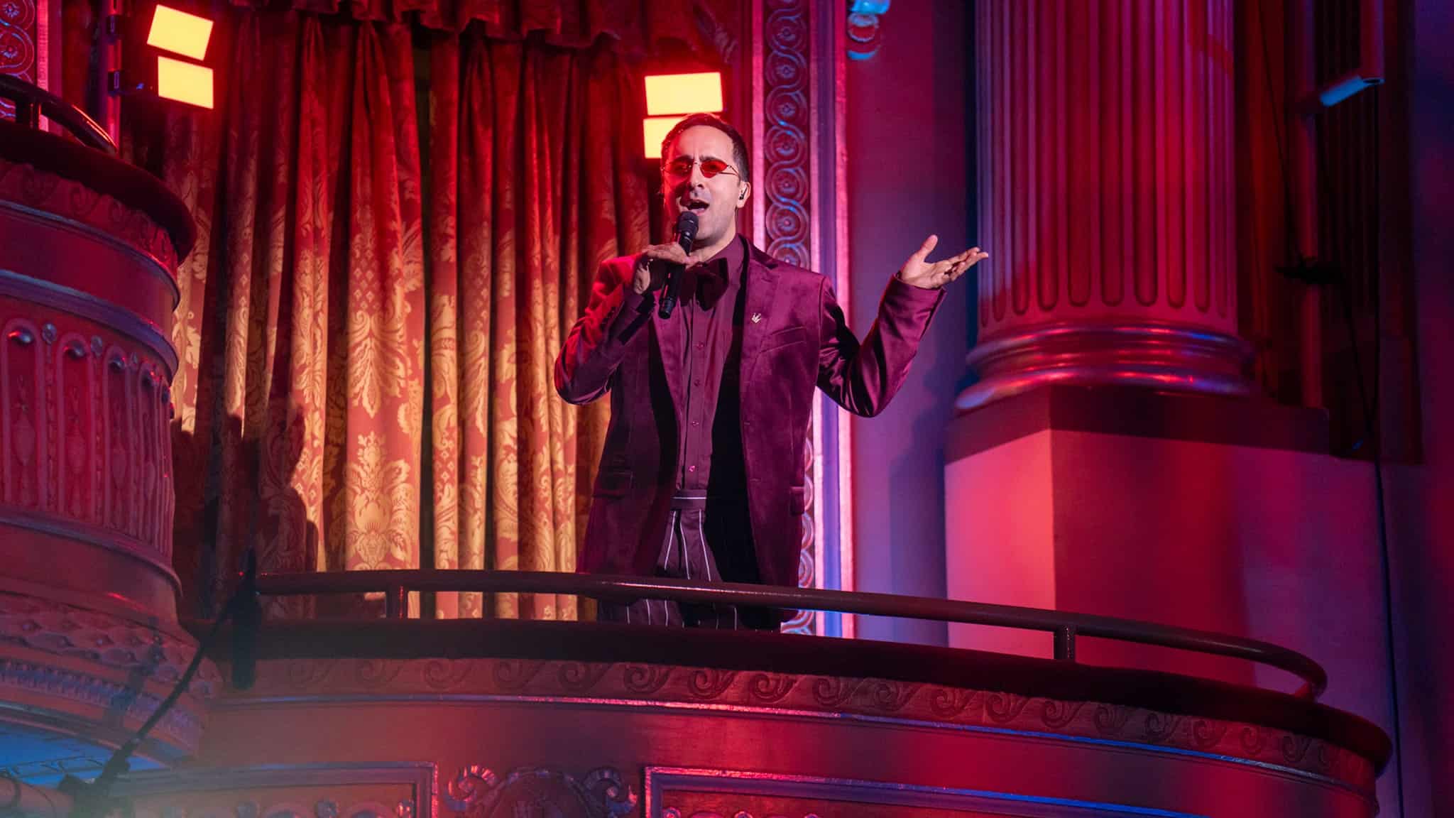 A person in a dark red suit sings or speaks into a microphone while standing on a balcony in a lavish, ornately decorated theater bathed in red and purple lighting.