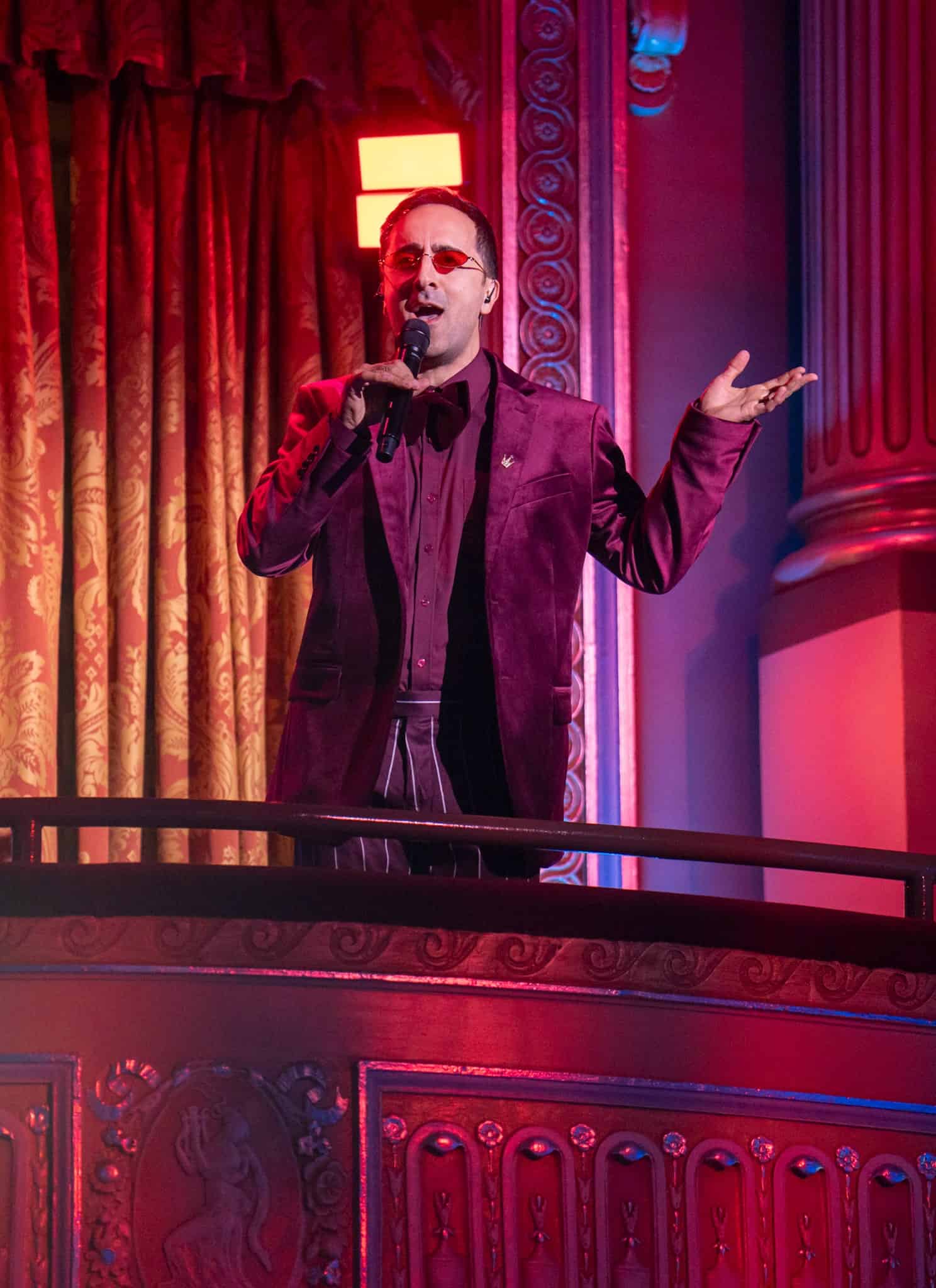 A person in a dark red suit sings or speaks into a microphone while standing on a balcony in a lavish, ornately decorated theater bathed in red and purple lighting.