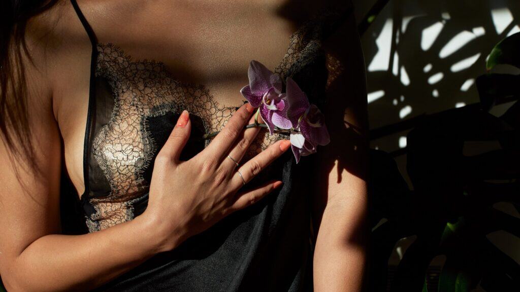 A woman wearing a sheer lace black camisole touches her chest with one hand, holding a purple orchid flower. Sunlight and shadows create patterns on her skin and clothing.