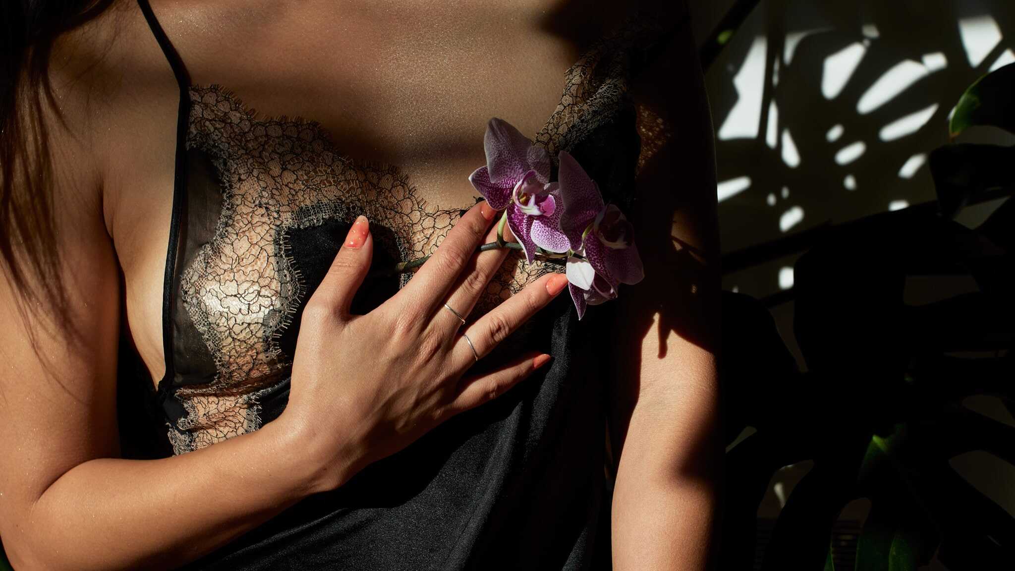 A woman wearing a sheer lace black camisole touches her chest with one hand, holding a purple orchid flower. Sunlight and shadows create patterns on her skin and clothing.