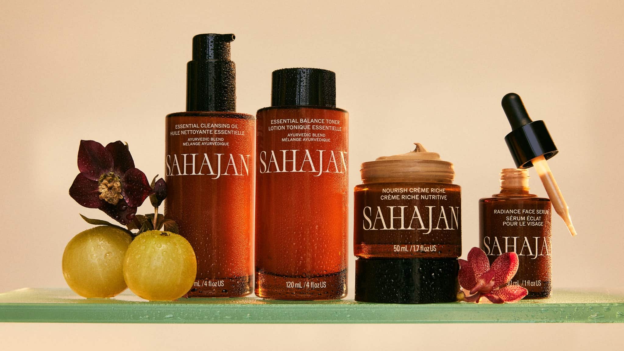 Four Sahajan skincare products are displayed with a flower, two amla fruits, and an orchid on a glass shelf against a beige background. One jar is open with cream exposed, and a dropper rests on another jar.