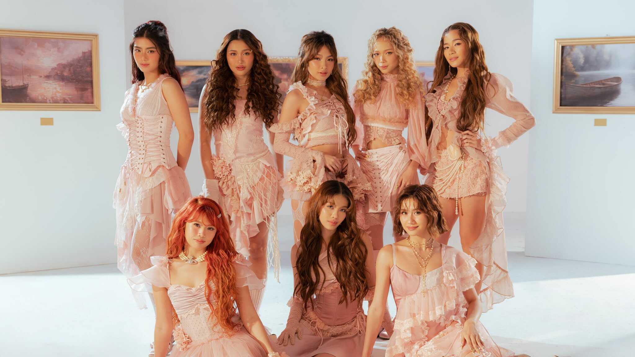 Eight women dressed in coordinated light pink, ruffled dresses pose together in a bright room with soft lighting and framed landscape paintings on the white walls behind them.