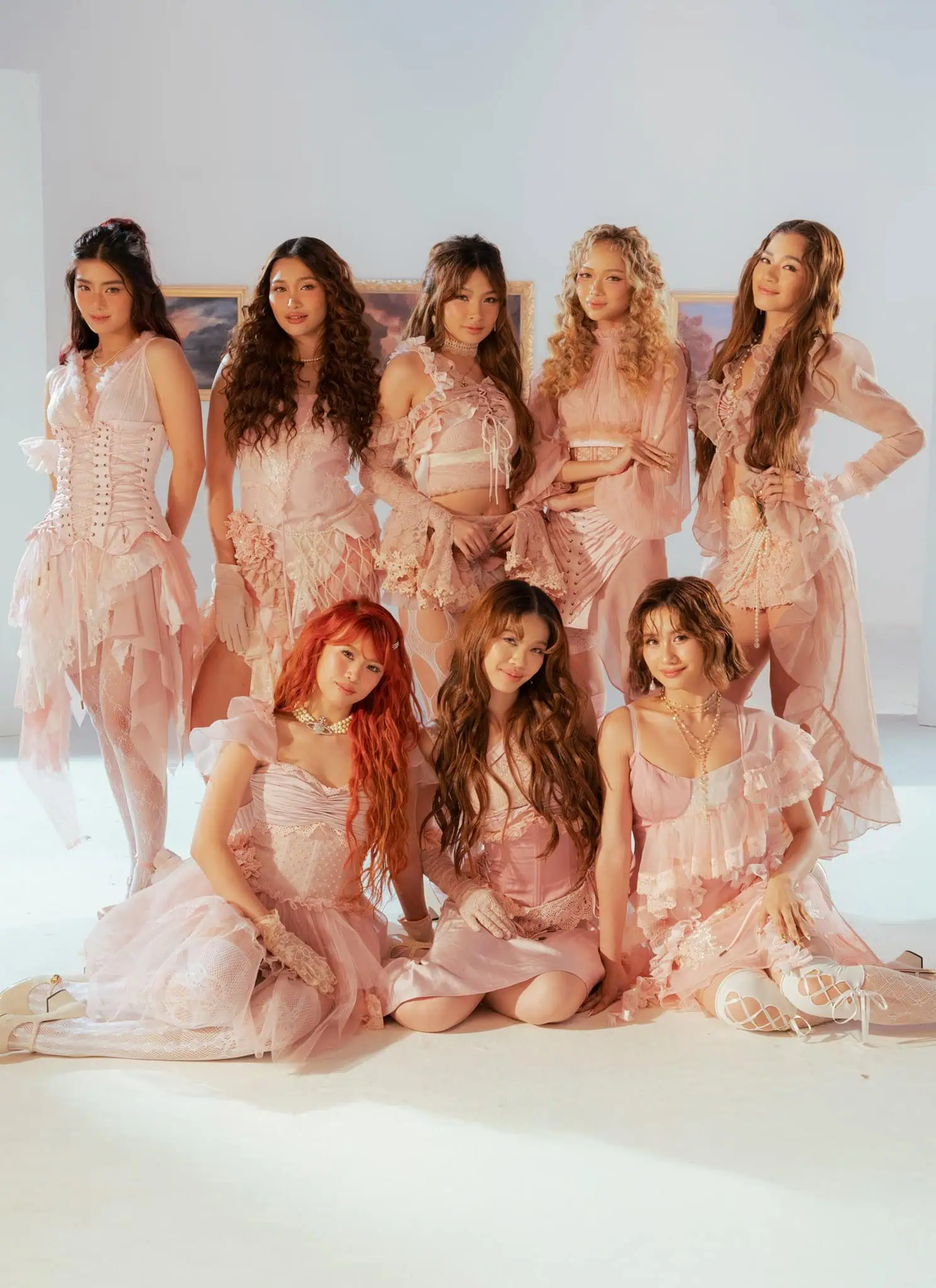 Eight women dressed in coordinated light pink, ruffled dresses pose together in a bright room with soft lighting and framed landscape paintings on the white walls behind them.
